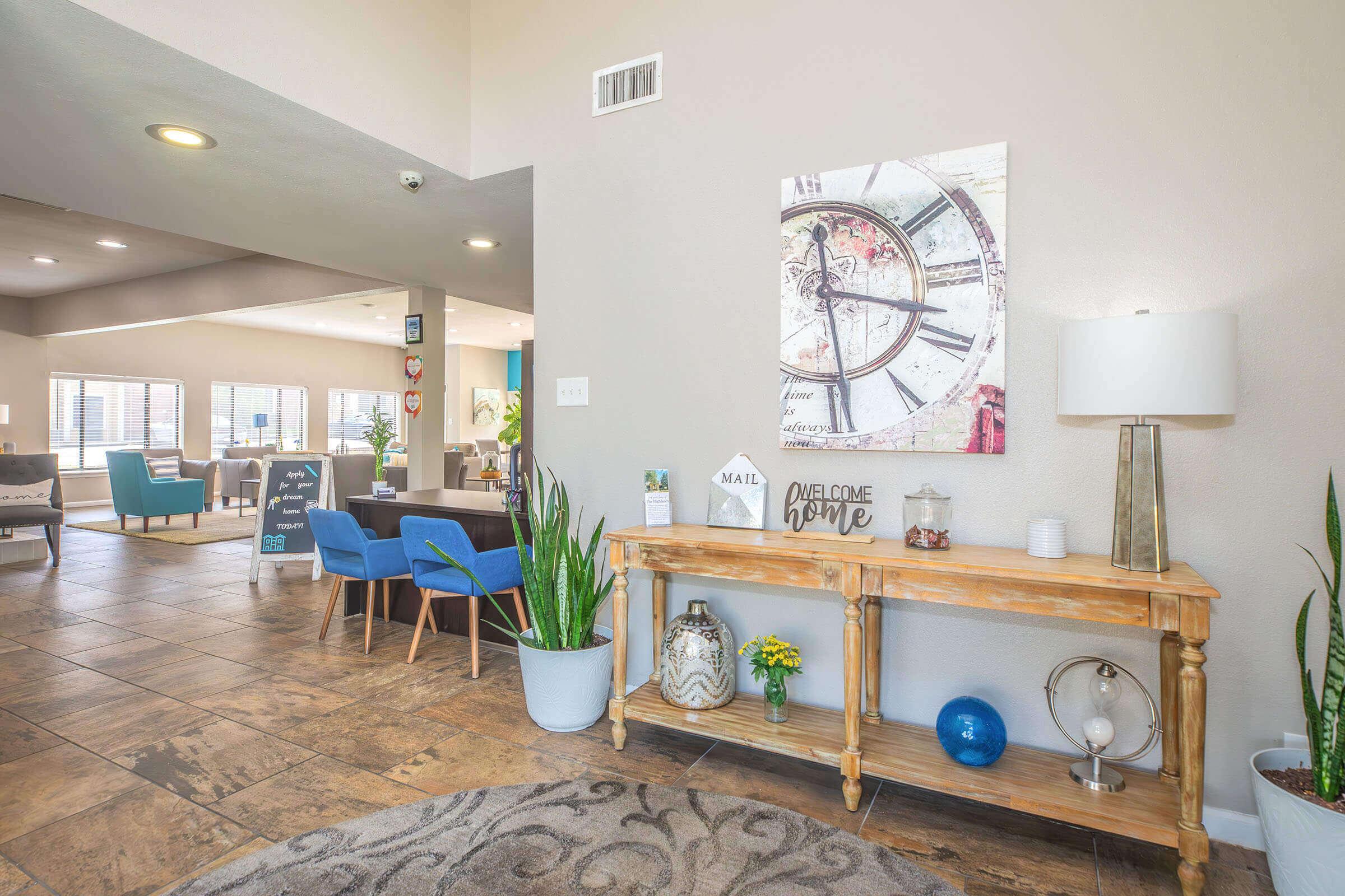 A welcoming lobby area featuring a wooden console table with decorative items, a lamp, and potted plants. There's artwork of a clock on the wall, and the space includes comfortable seating arrangements with blue chairs. The setting is bright and inviting, with tile flooring and natural light.