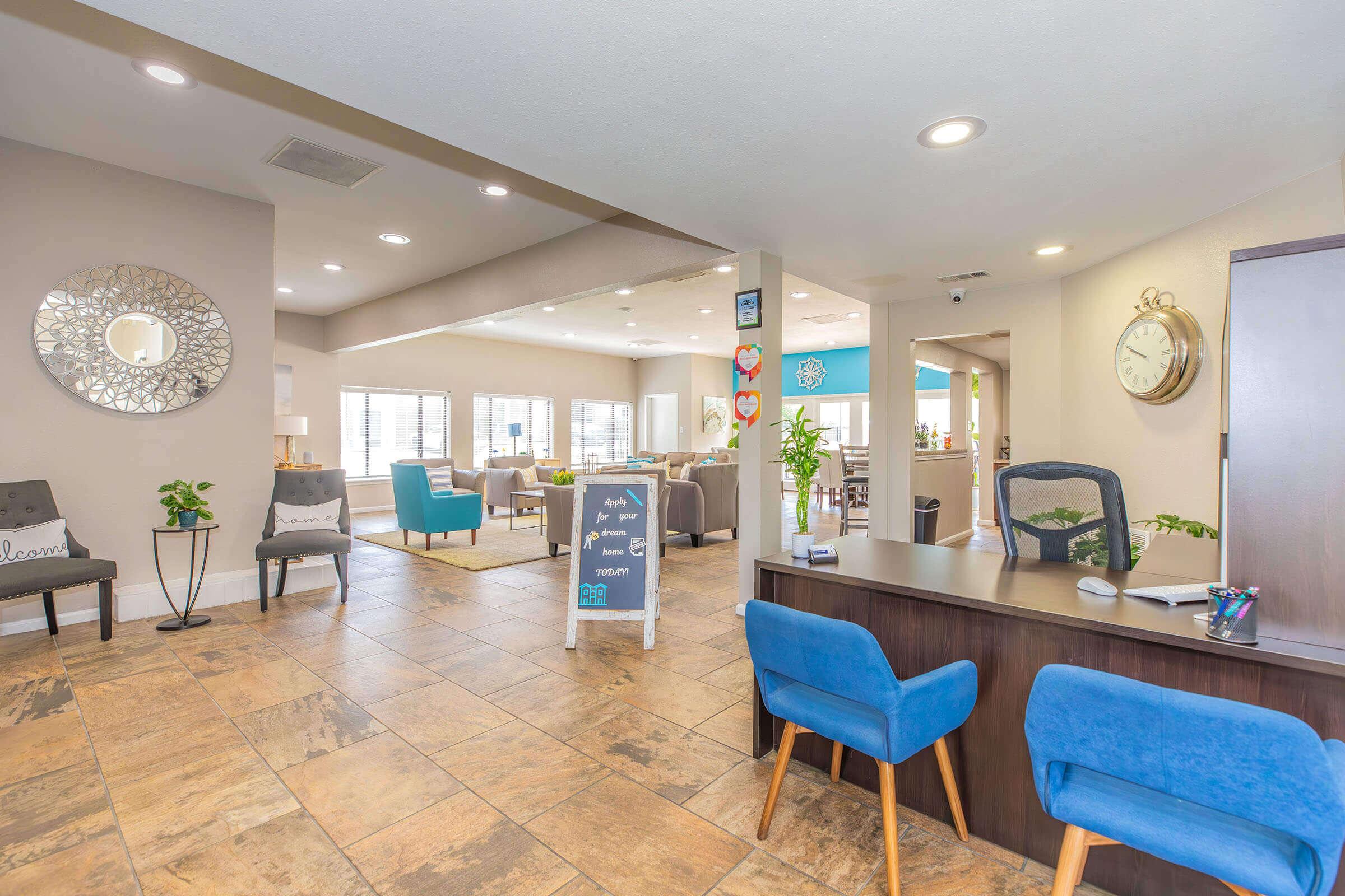 A bright and modern reception area featuring a desk with blue chairs, decorative plants, and a stylish wall mirror. The space includes a cozy seating area with light-colored furniture and large windows, creating an inviting atmosphere. A signboard with a message is placed near the desk.
