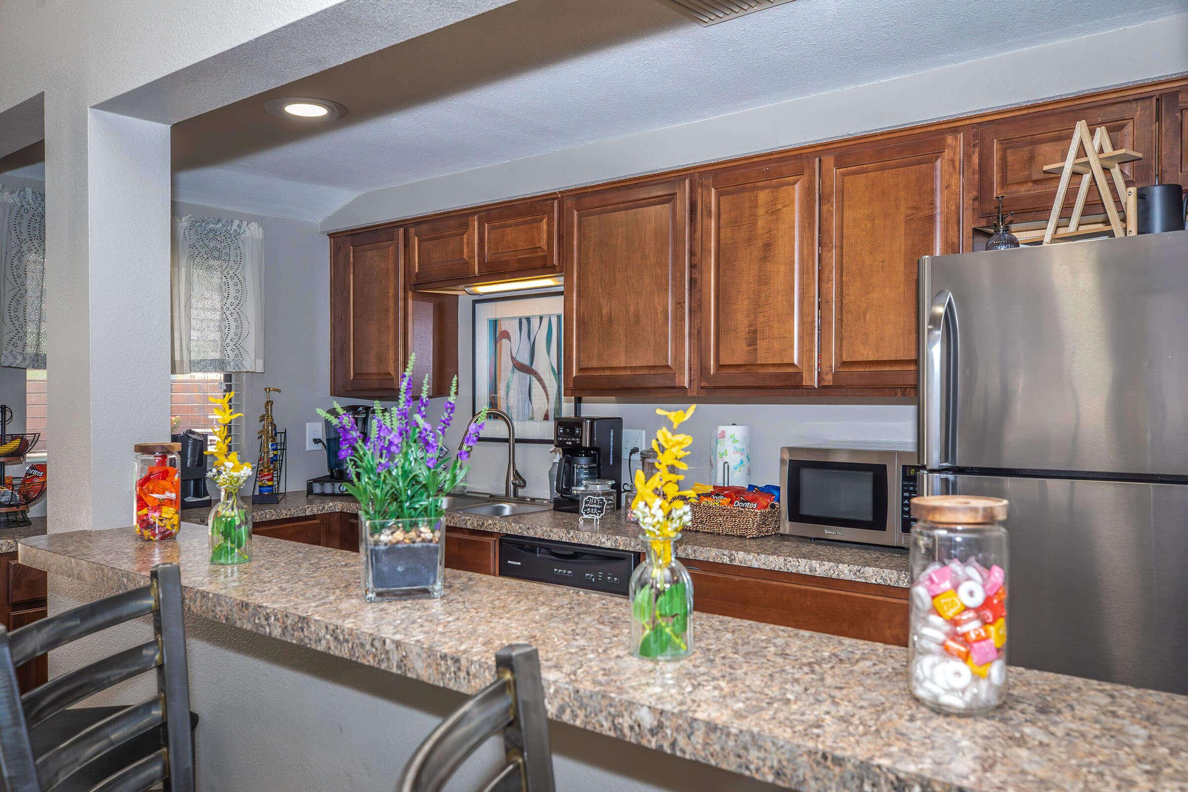 A modern kitchen featuring wooden cabinets, stainless steel appliances, and a countertop with small vases containing flowers. There’s a coffee maker, microwave, and colorful snacks visible on the counter, creating a welcoming and functional space.