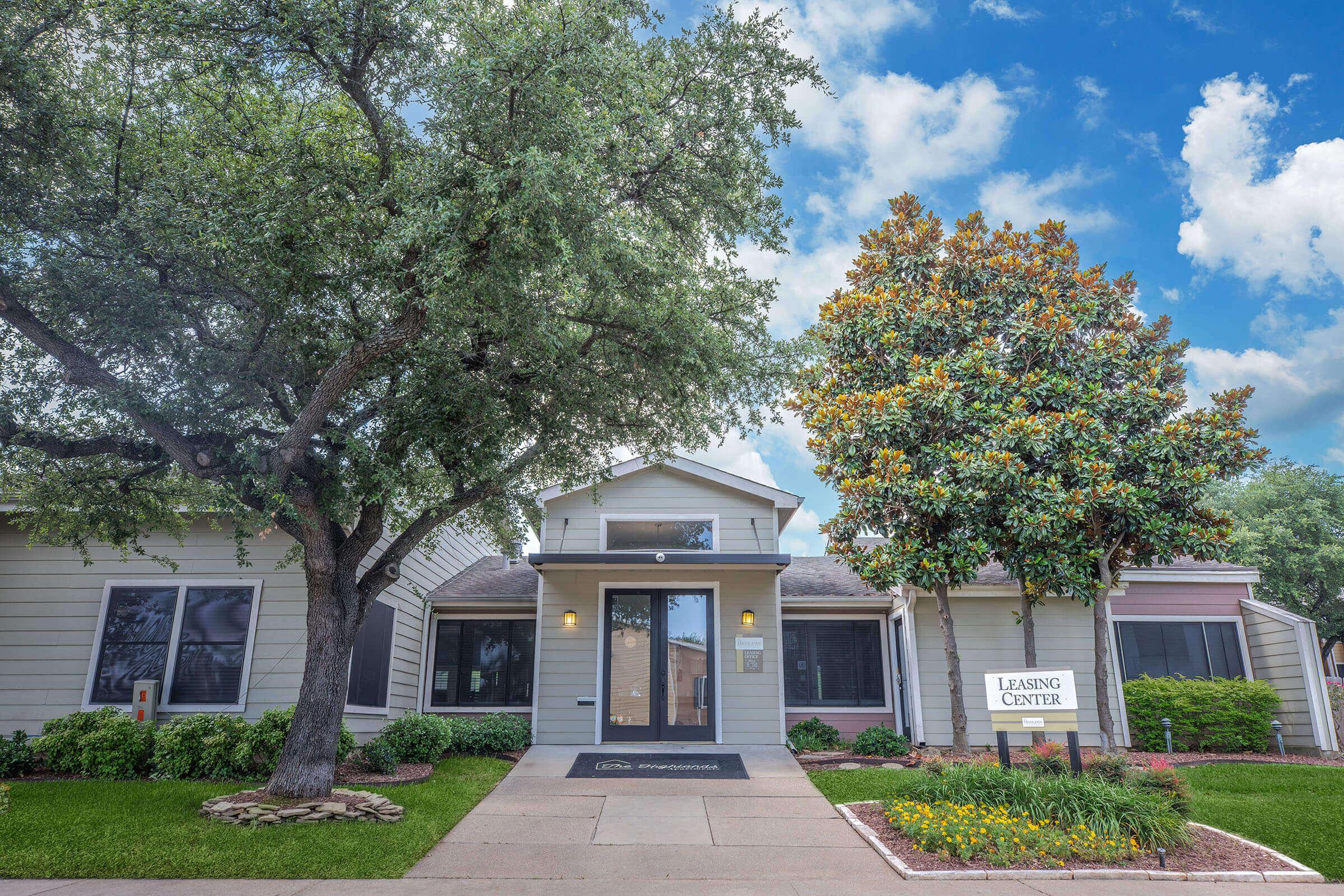 A charming leasing center with a welcoming entrance, framed by lush trees and well-maintained landscaping. The building features a light-colored exterior, a central door, and a sign that reads "Leasing Center." The sky is partly cloudy, adding to the pleasant atmosphere.