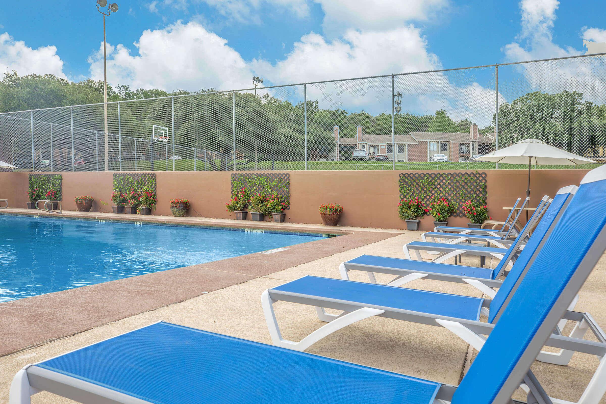 A serene swimming pool area featuring bright blue lounge chairs arranged around the pool. Lush greenery and flower pots adorn the edges, while a basketball hoop is visible in the background. The sky is partly cloudy, adding to the inviting atmosphere of the outdoor space.