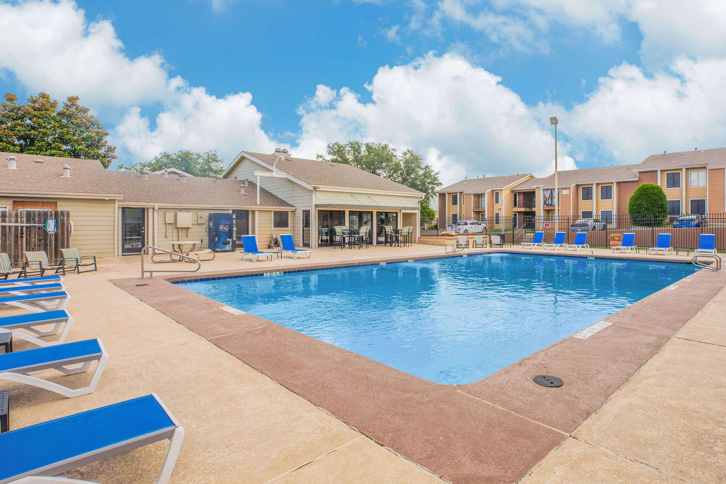 A clear blue swimming pool surrounded by lounge chairs, with a nearby building featuring large windows. Lush green trees are visible in the background, and the sky is partly cloudy. The area appears inviting and well-maintained, ideal for relaxation and leisure activities.