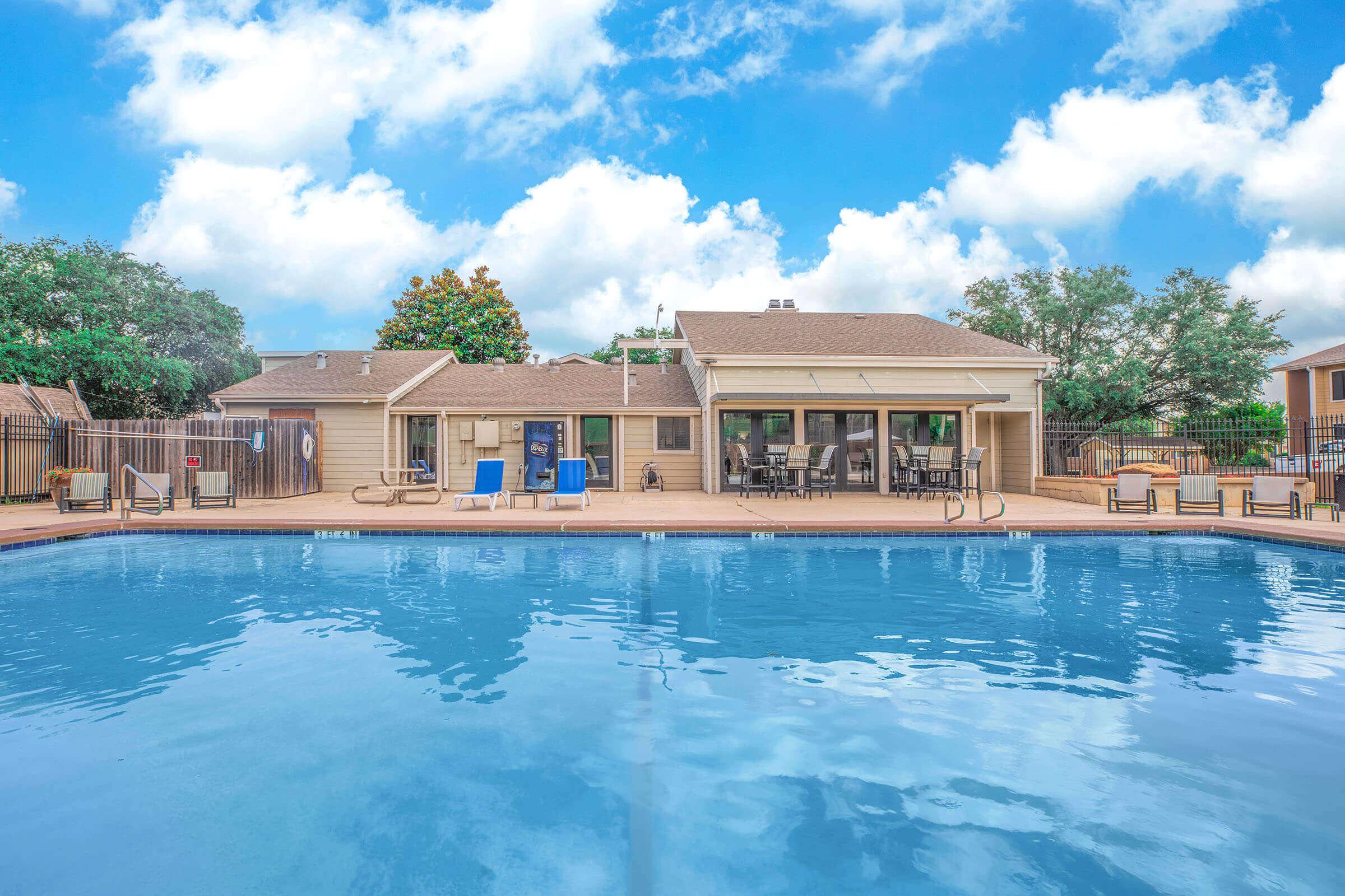 A clear blue swimming pool surrounded by a patio with lounge chairs and tables. In the background, a residential building with large windows and a green landscape. Fluffy clouds and a bright blue sky enhance the inviting atmosphere of the area.