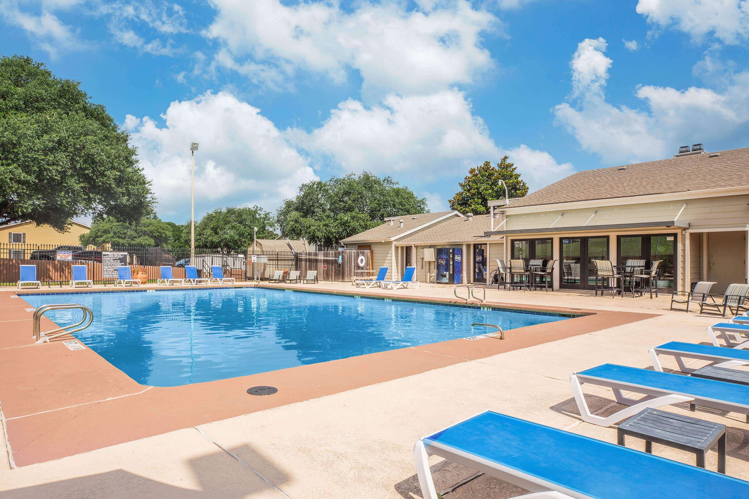 A clear blue swimming pool surrounded by lounge chairs and a concrete deck. In the background, there's a building with large windows and outdoor seating, framed by green trees under a bright blue sky with fluffy white clouds.