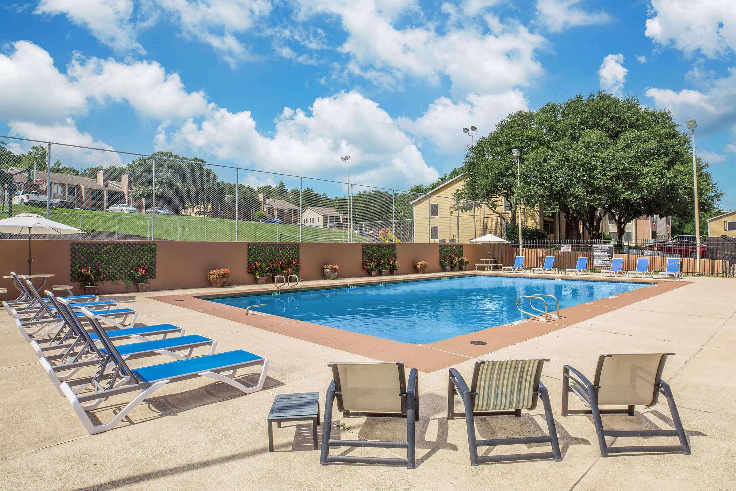 A sunny outdoor swimming pool area with lounge chairs arranged around the pool. Surrounding greenery and tall trees provide shade, while umbrellas are set up for shade. A fence encloses the pool area, and a few residential buildings are visible in the background against a blue sky with scattered clouds.
