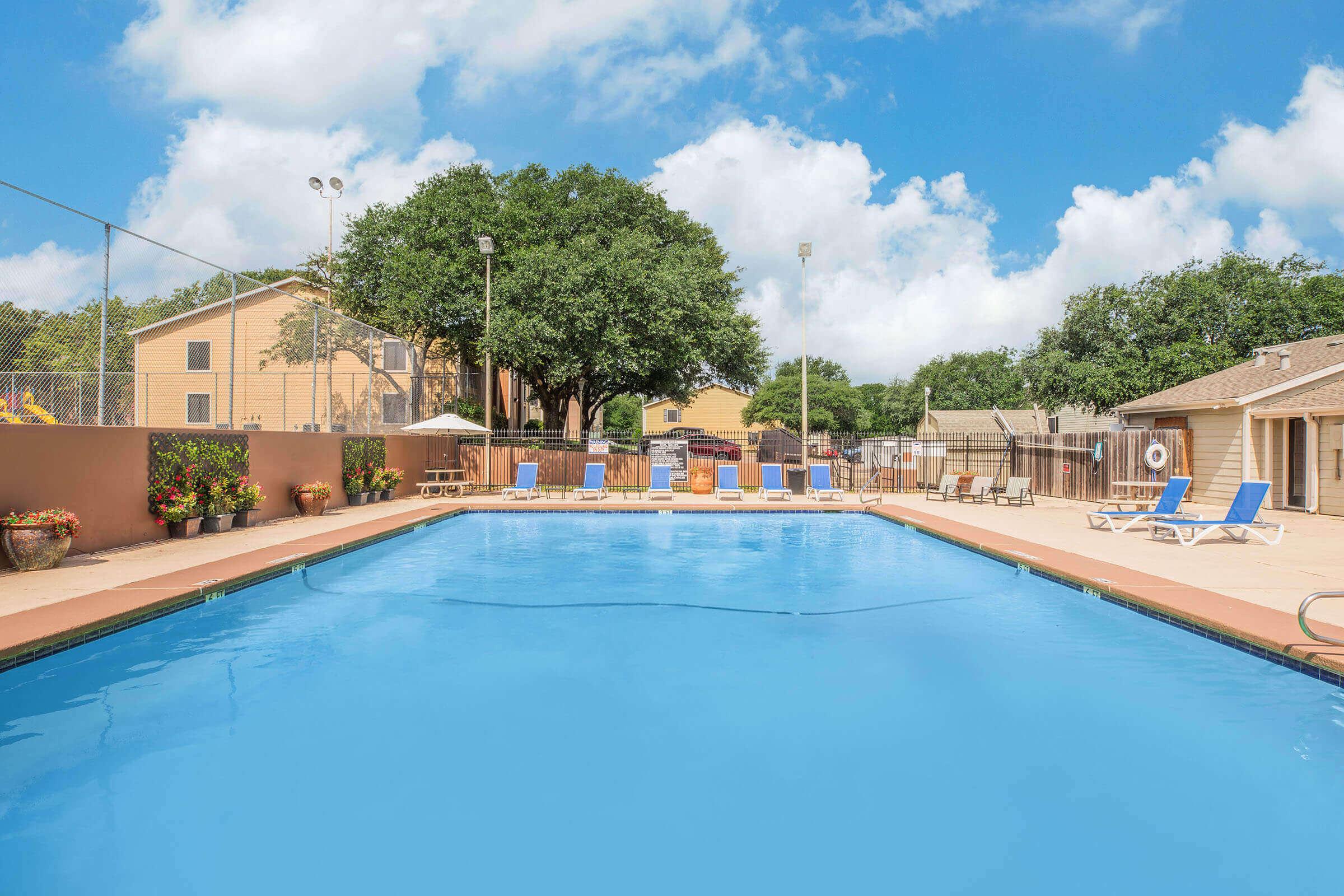A sunny outdoor pool area featuring a vibrant blue swimming pool surrounded by lounge chairs. Lush green trees and bright skies with fluffy clouds provide a pleasant backdrop. The setting includes decorative pots and fencing, creating a welcoming atmosphere for relaxation and leisure.