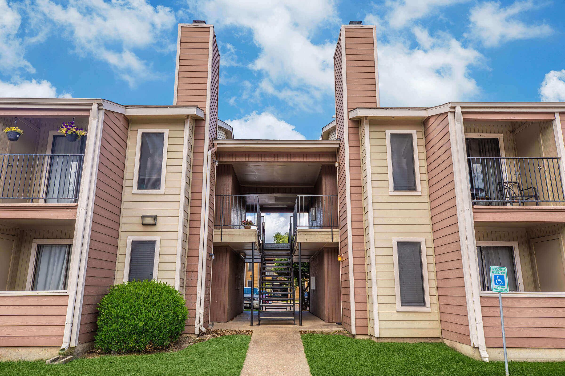Two-story apartment building with a central staircase leading to upper level apartments. The exterior features beige paneling with orange trim and small balconies. Lush green grass and a bush are in the foreground, under a bright blue sky with fluffy white clouds.