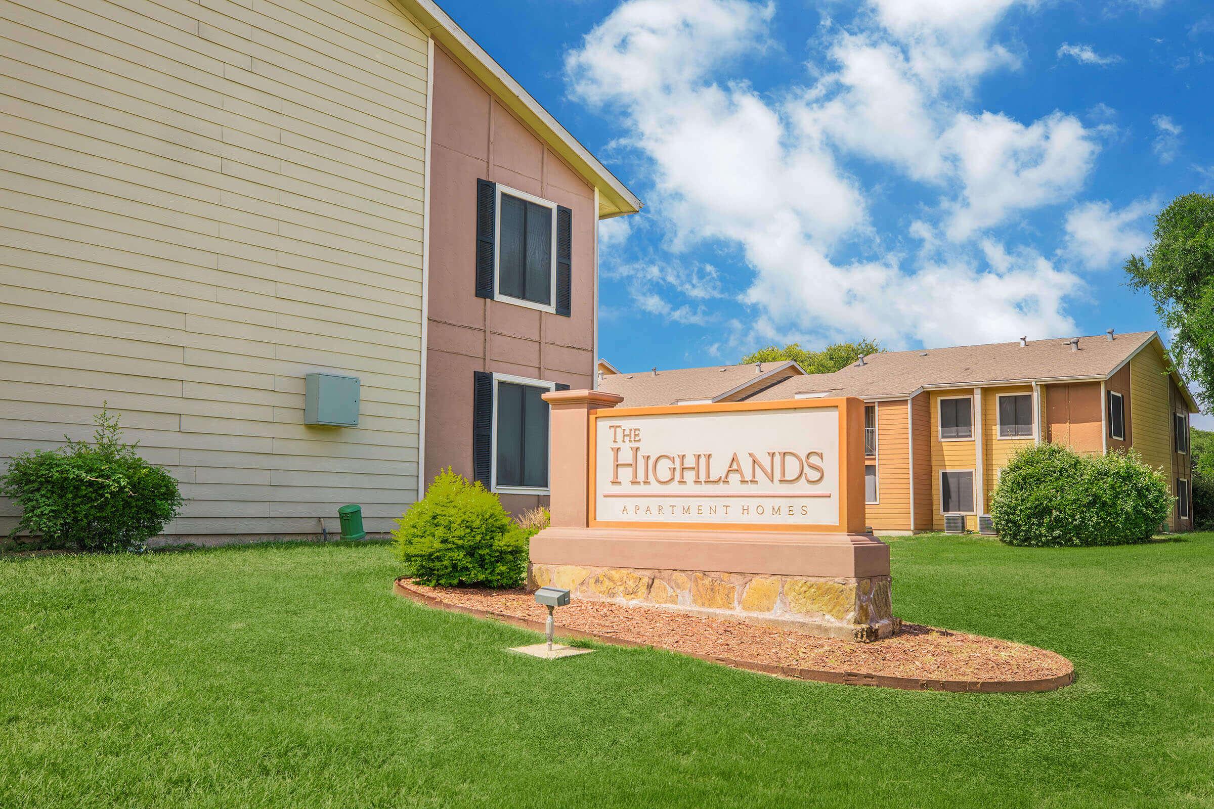 A sign for "The Highlands Apartment Homes" is prominently displayed in front of a well-maintained residential building. The landscape features green grass and shrubs, with a bright blue sky and fluffy clouds in the background. The buildings are two-storied with multiple windows.