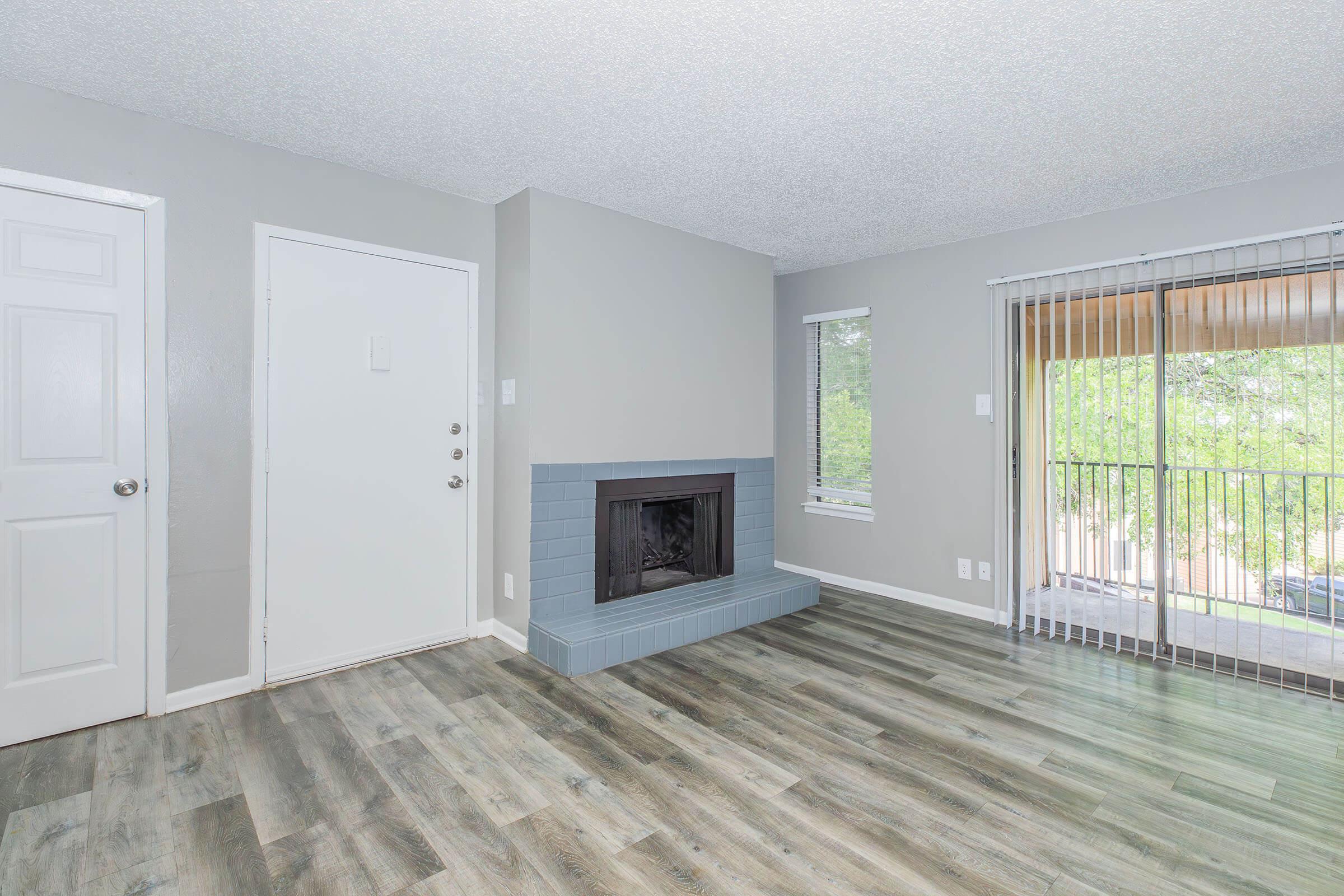 A modern living room featuring light gray walls, a blue tiled fireplace, and large windows allowing natural light. The floor is covered with laminate wood flooring, and a sliding glass door leads to a balcony. A white door on the left provides access to another room or entrance.