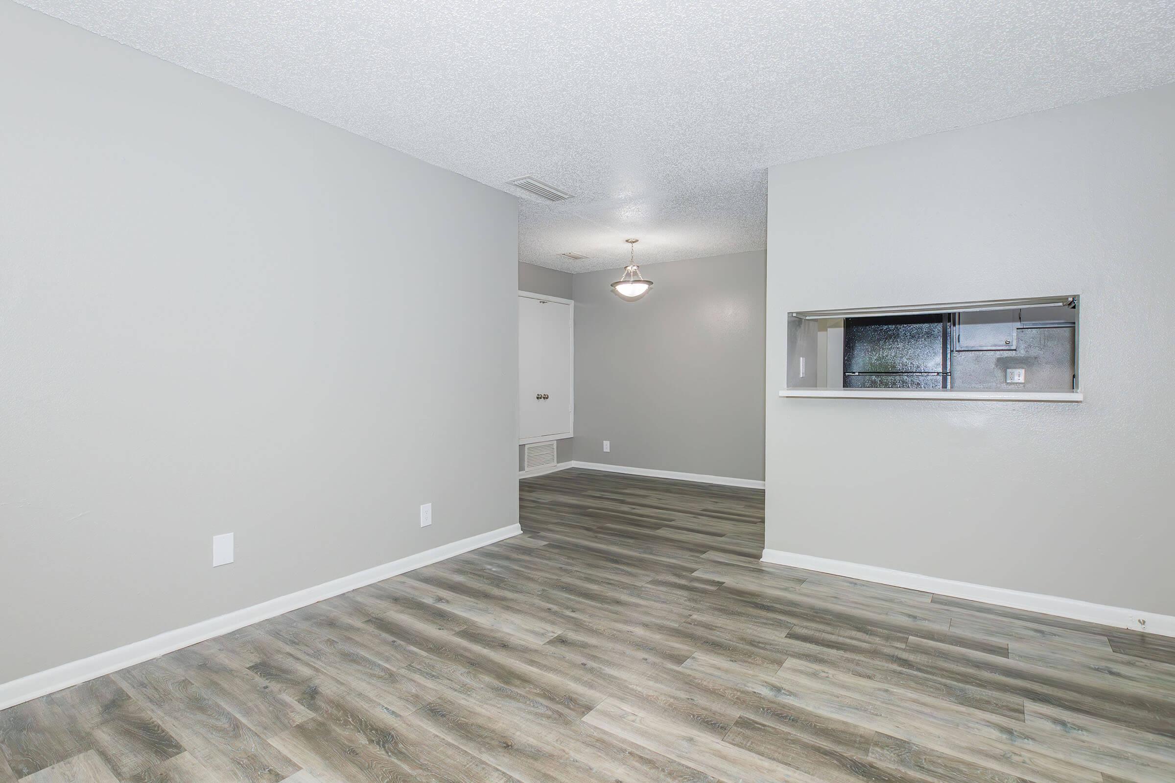 A spacious, empty living area with light gray walls and modern vinyl flooring. A small dining area is visible through a passthrough window, with a ceiling light fixture hanging above. The room is well-lit and features no furniture, offering a clean, open layout.
