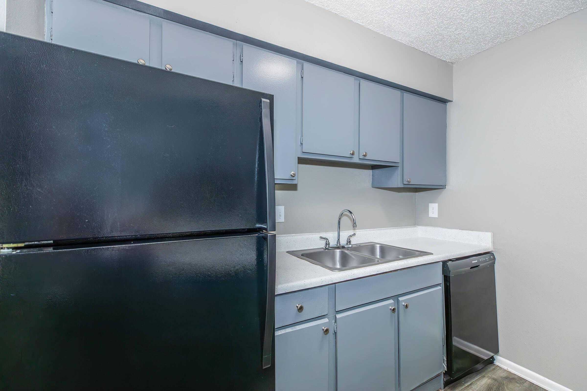 A compact kitchen with dark blue cabinets, a black refrigerator, a stainless steel dishwasher, and a double sink with a modern faucet. The countertops are light-colored, and the walls are painted a neutral gray, creating a clean and functional cooking space.