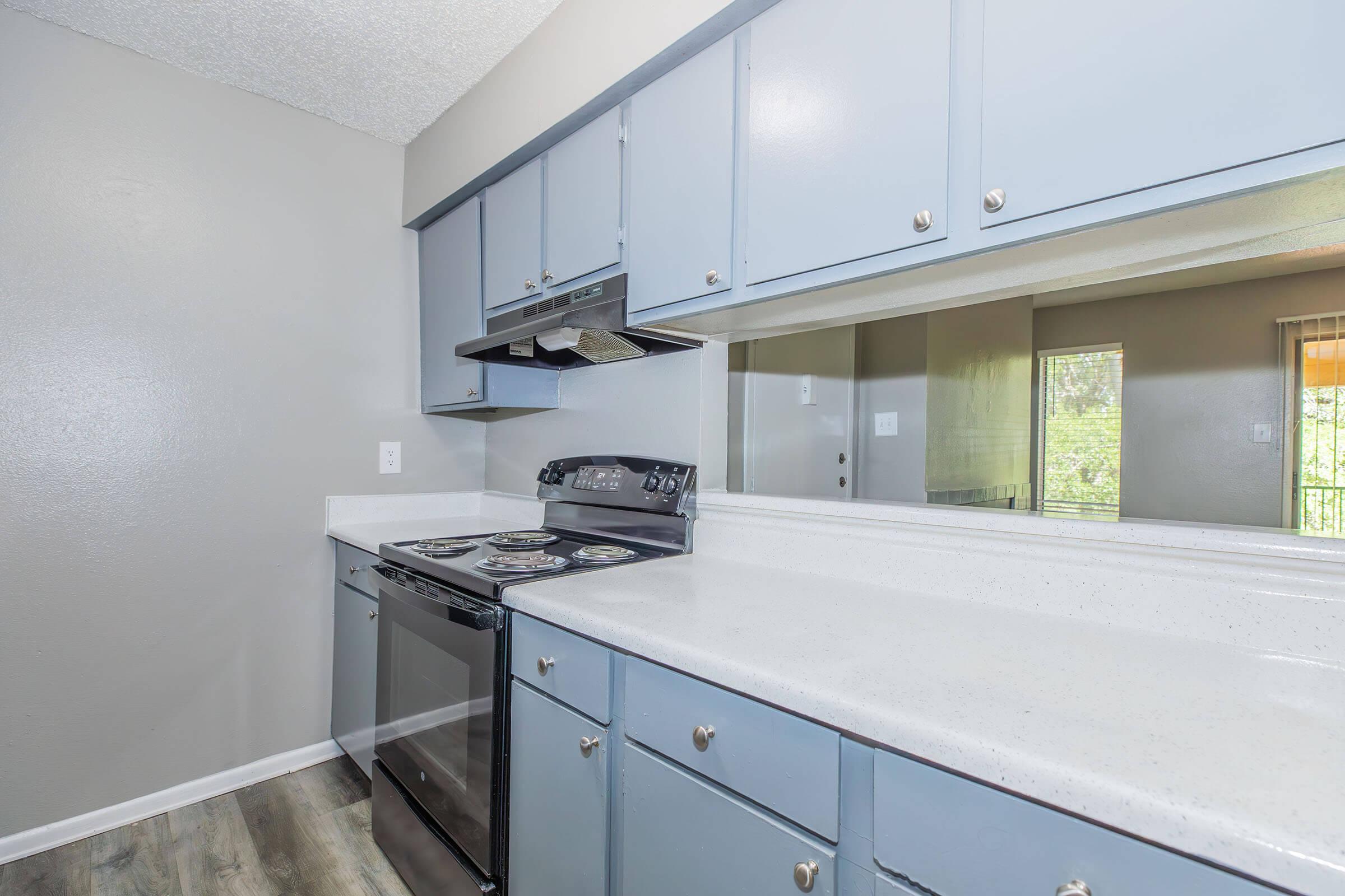 A modern kitchen featuring light blue cabinetry, a black stove, and a sleek countertop. The space has an open layout with a mirror reflecting a bright area. The flooring is a neutral color that complements the walls and cabinets, creating a clean and inviting atmosphere.