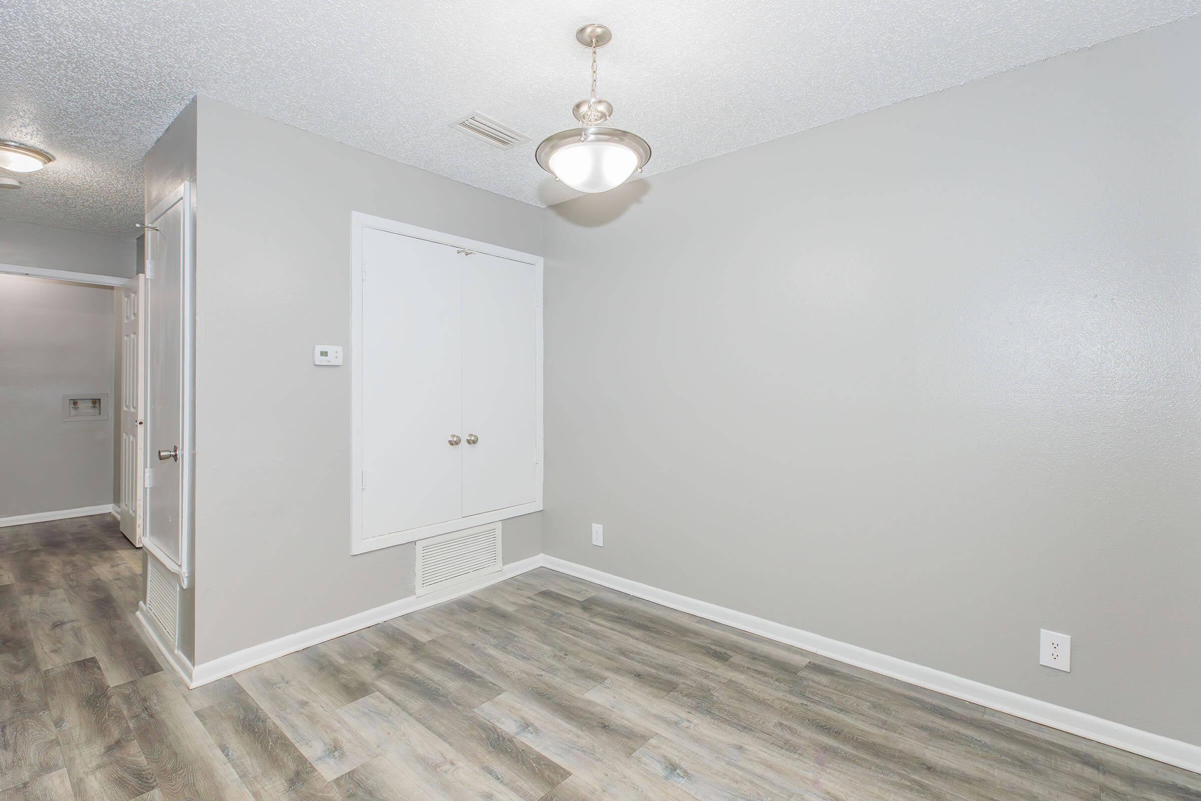 Interior view of a room featuring gray walls, a light fixture hanging from the ceiling, and a closet with double doors. The floor is covered in light-colored laminate wood. A vent is visible on the wall, and there is a hallway leading to another area in the background.