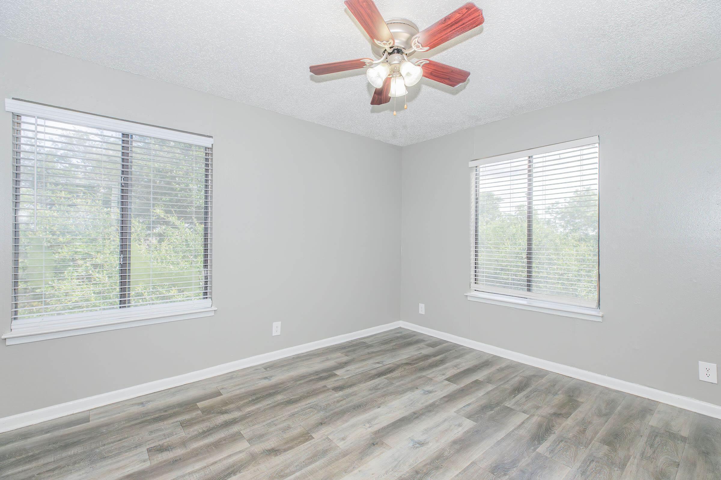 Empty room with gray walls and two windows featuring white blinds. A ceiling fan with wooden blades hangs in the center. The floor has light-colored planks, creating a clean and modern look. greenery is visible outside through the windows, adding a touch of nature.