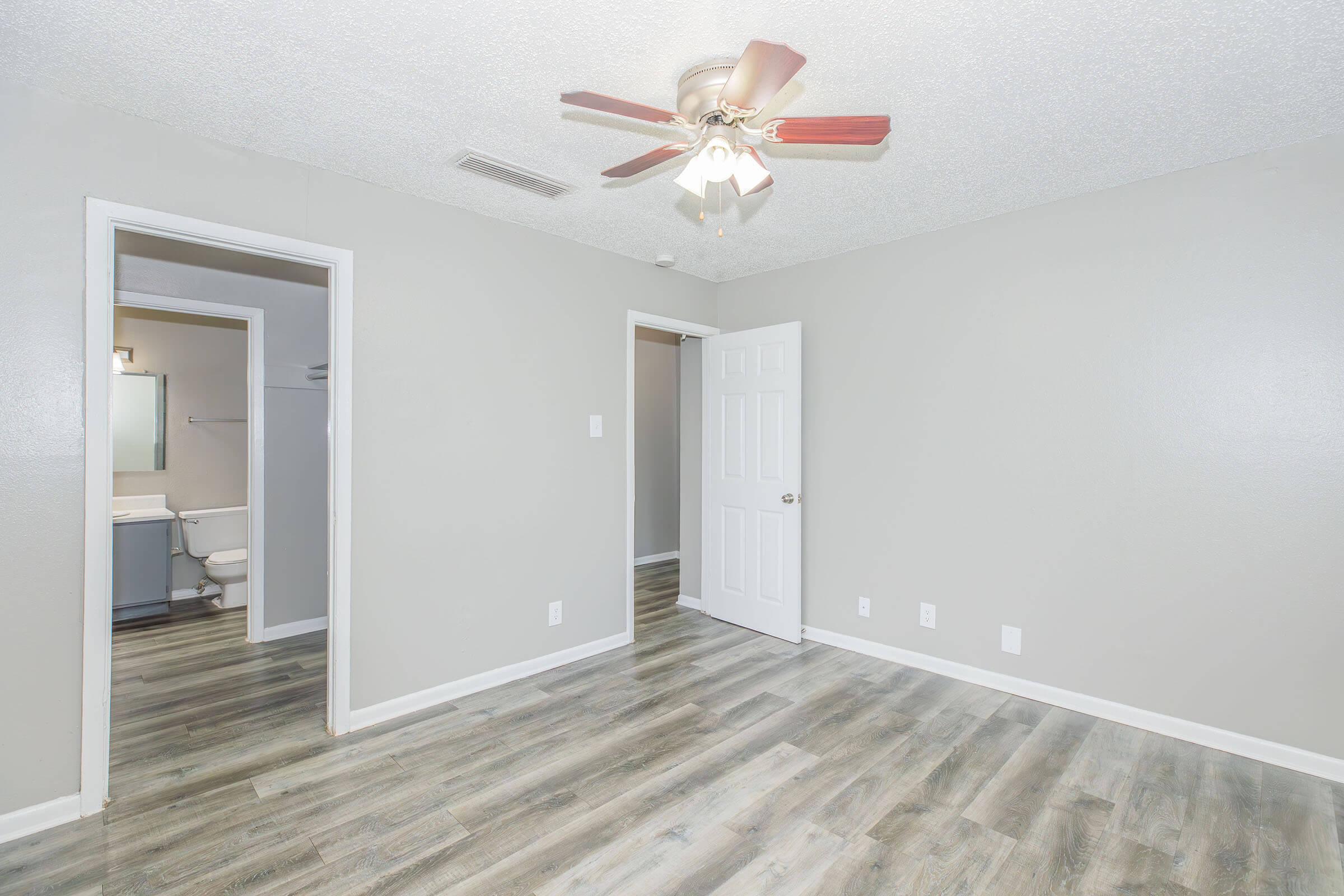 A spacious room with a ceiling fan, light gray walls, and light wood-like flooring. There is a doorway leading to a bathroom visible in the background, with a white door closed to the left. The room appears bright and modern, showcasing clean lines and neutral decor.