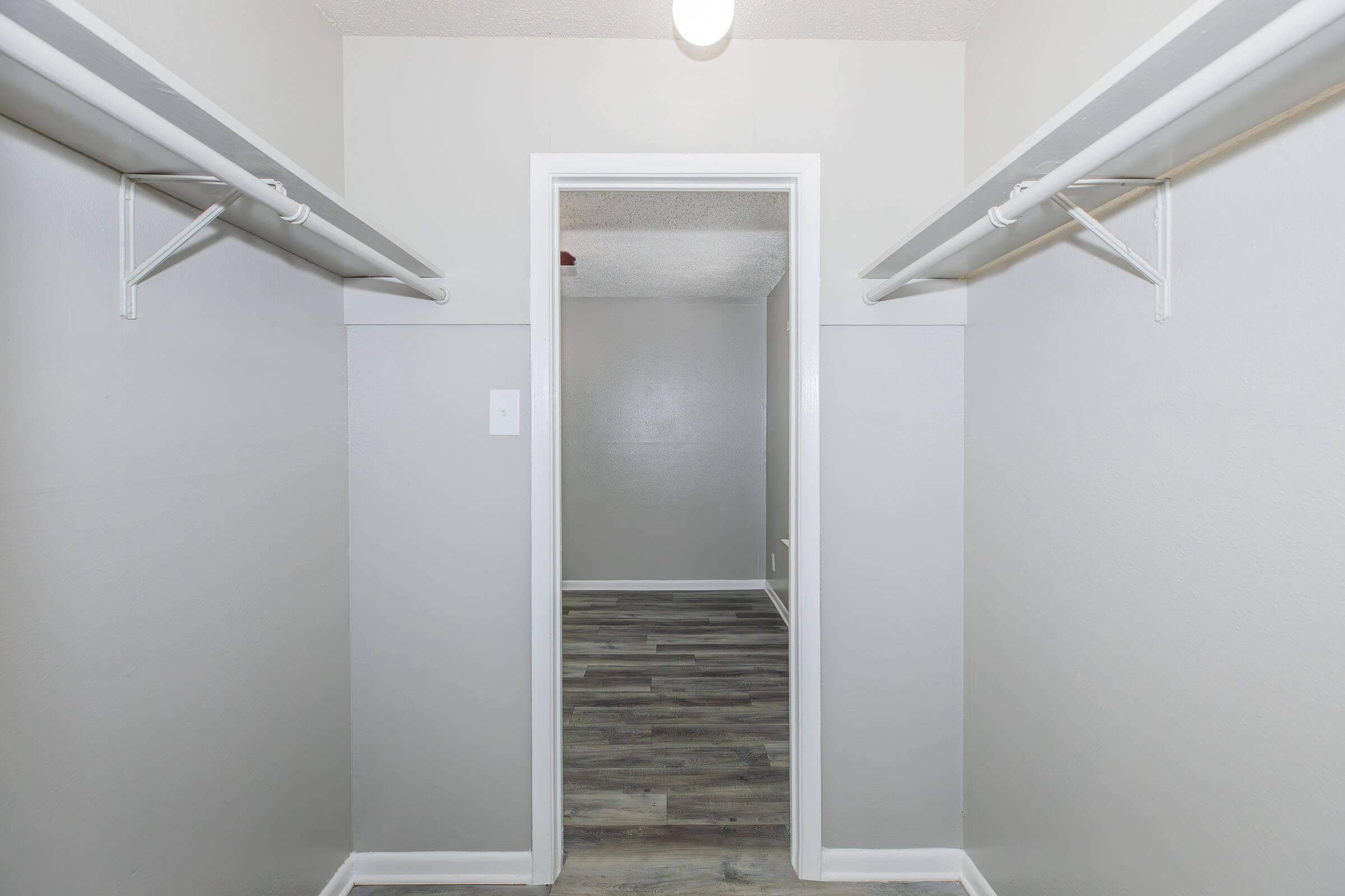 A clean and spacious hallway with light gray walls, featuring two open closets on either side. The floor is finished in a wood-like pattern, and a light fixture is visible on the ceiling. A doorway at the end leads to another room.
