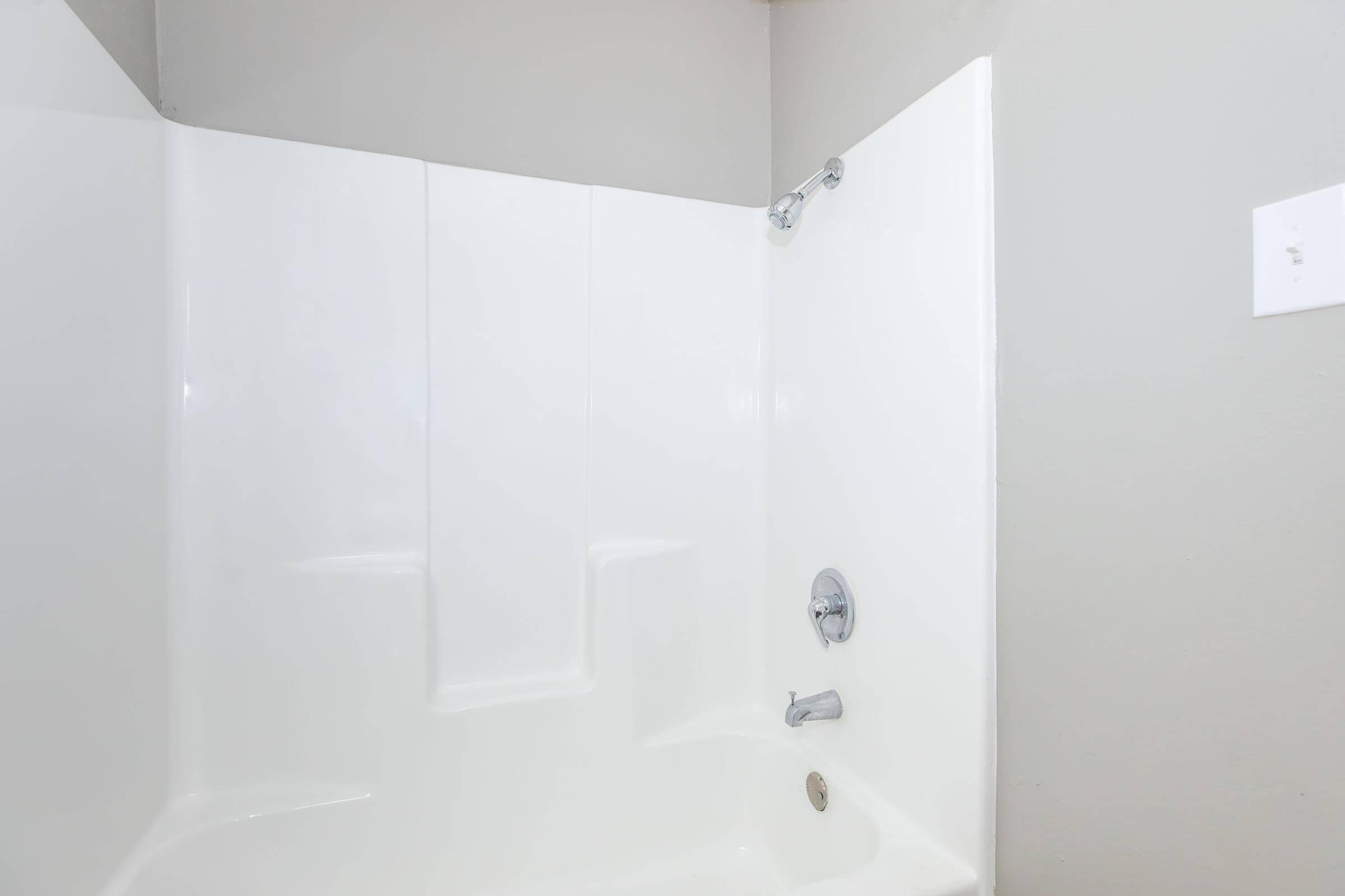 A clean, modern shower stall with white acrylic walls and a chrome showerhead. The shower has a built-in shelf for toiletries and features a single control knob for temperature. The surrounding walls are painted in a light gray color, creating a bright and airy atmosphere.