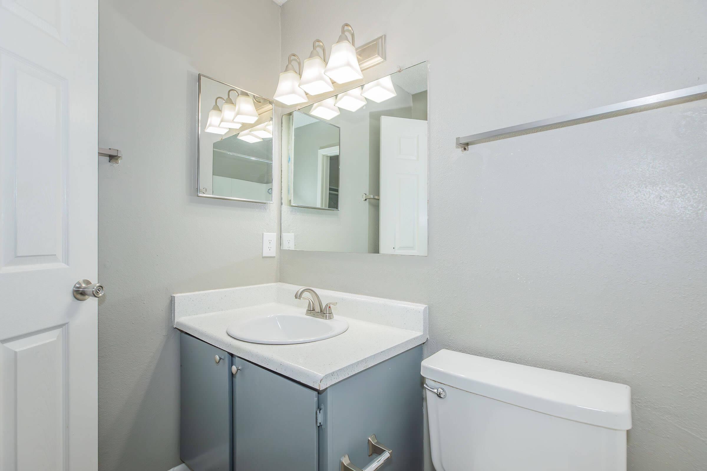 A clean and modern bathroom featuring a white sink with a chrome faucet, a large mirror, three light fixtures above, and gray walls. There’s a blue cabinet beneath the sink and a white toilet adjacent to it. A towel bar is mounted on the wall. The overall aesthetic is sleek and well-lit.