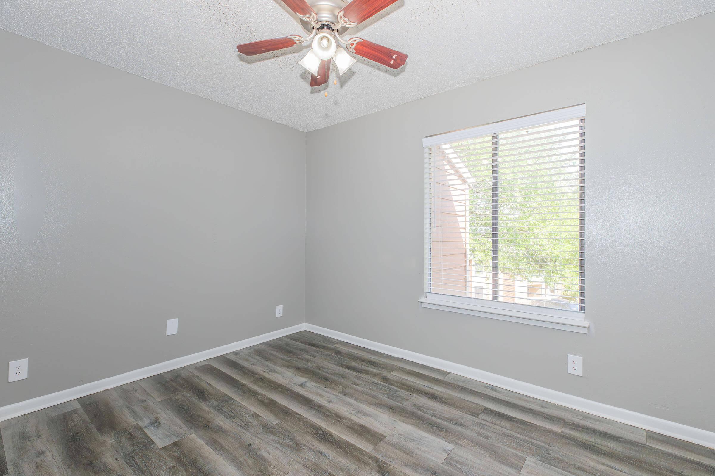 Empty room with light gray walls and a ceiling fan with wooden blades. Natural light filters through a closed window with blinds. The floor features wood-like laminate in a warm tone. No furniture or decorations are present.