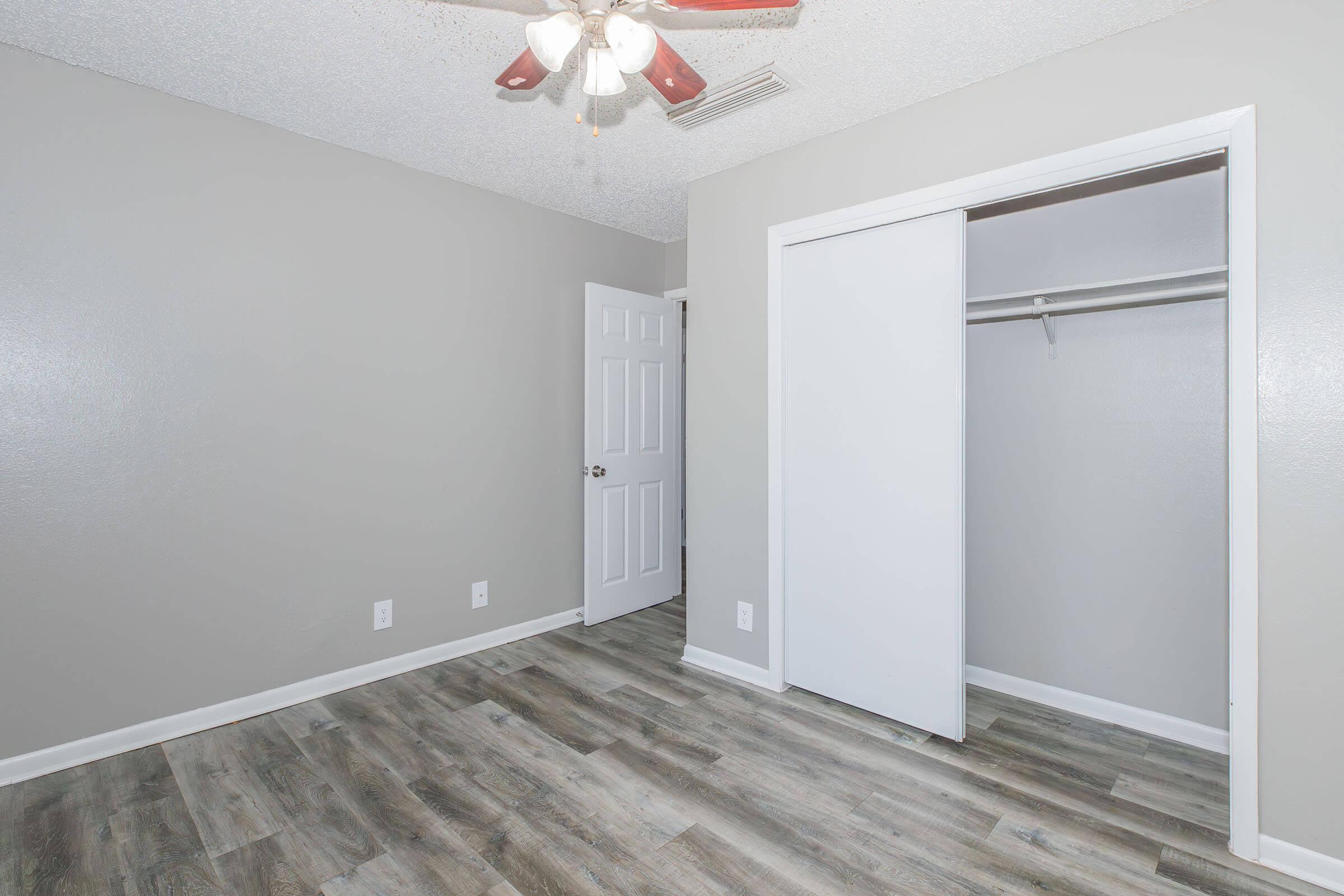A small, unfurnished room with a light gray wall and a ceiling fan. The flooring is wood-like, and there is a white closet with sliding doors. A white door leads to another area, and there are no decorations or furniture in the space, giving it a clean, minimalist appearance.