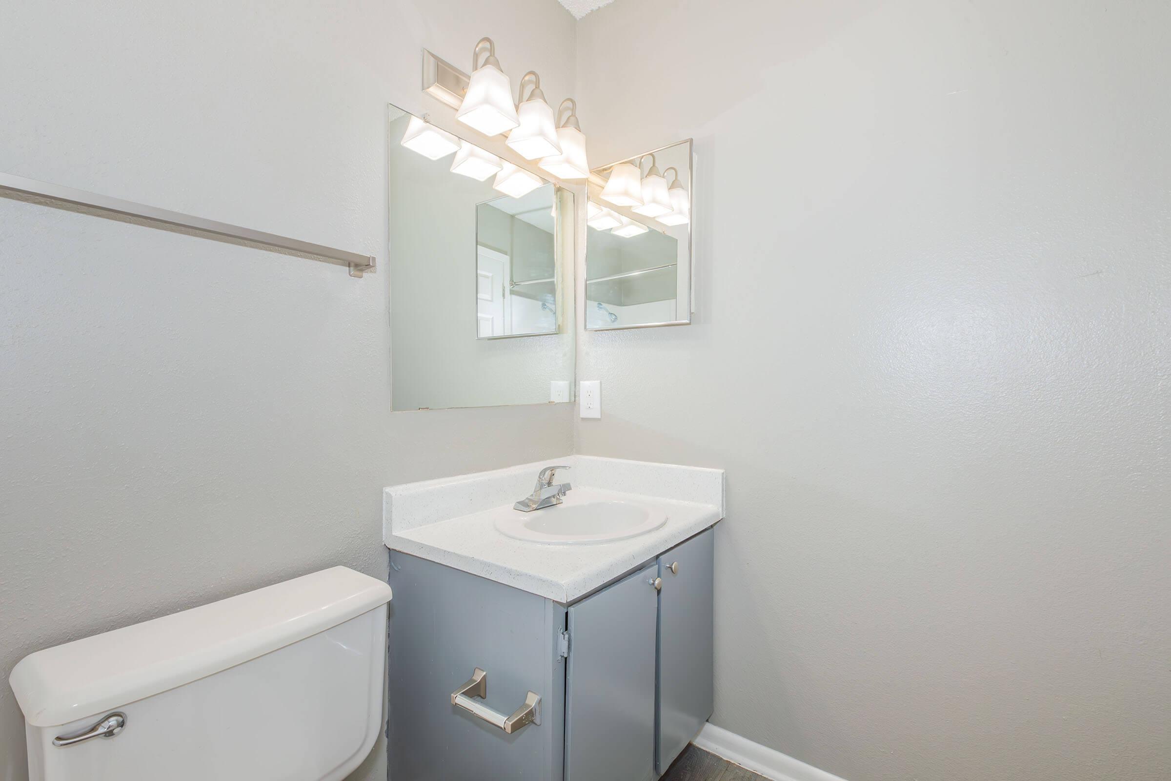 A modern bathroom featuring a white sink with a gray cabinet, a toilet, and a mirror with five light fixtures above. The walls are painted in a light gray color, and the floor is dark. The overall design is clean and contemporary.