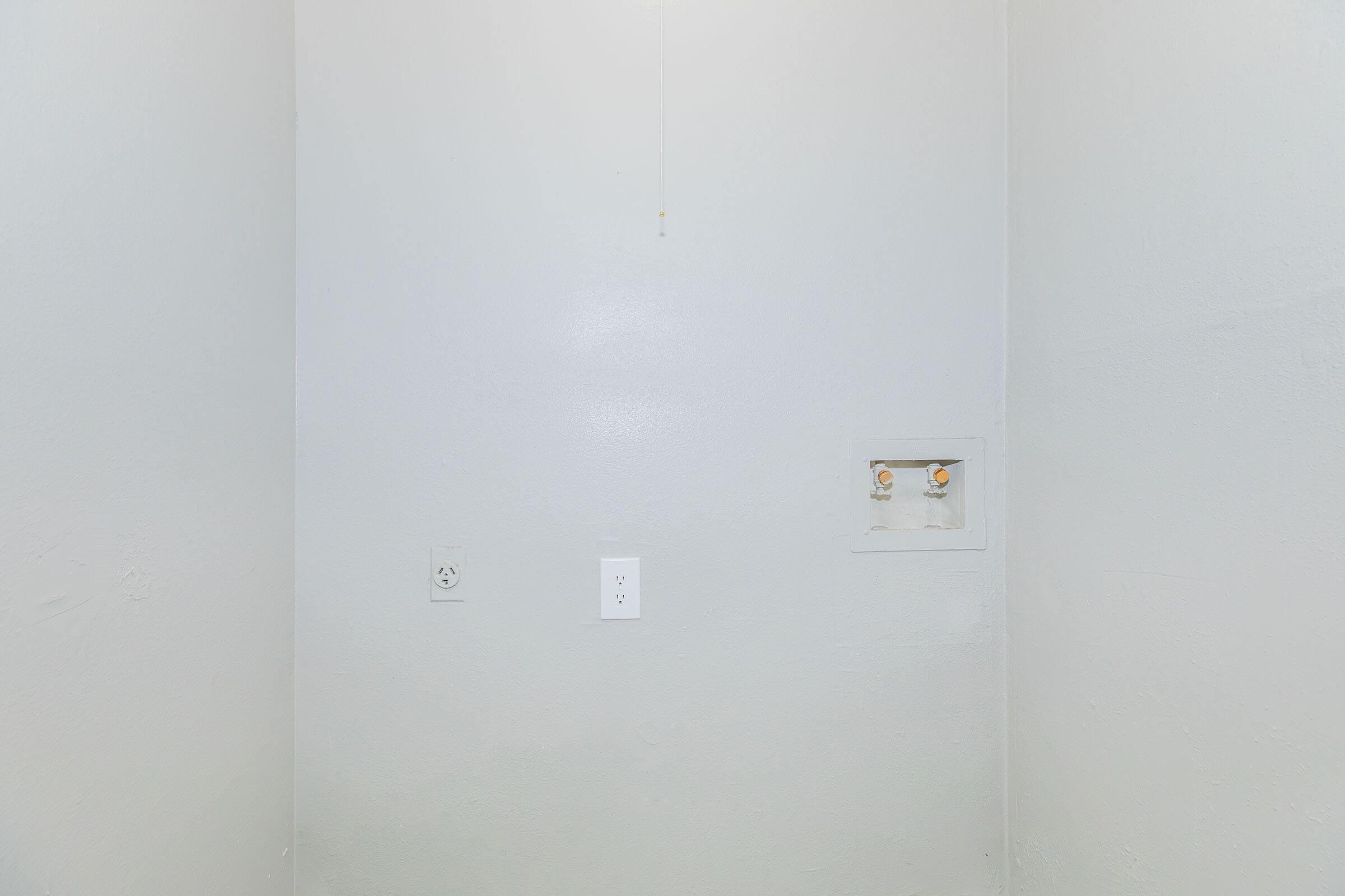 A plain, light gray wall with an electrical outlet and a switch. To the right, there are two water connections visible in a small recessed area. The overall appearance is minimalistic and simple, with no decorative elements or furniture present.