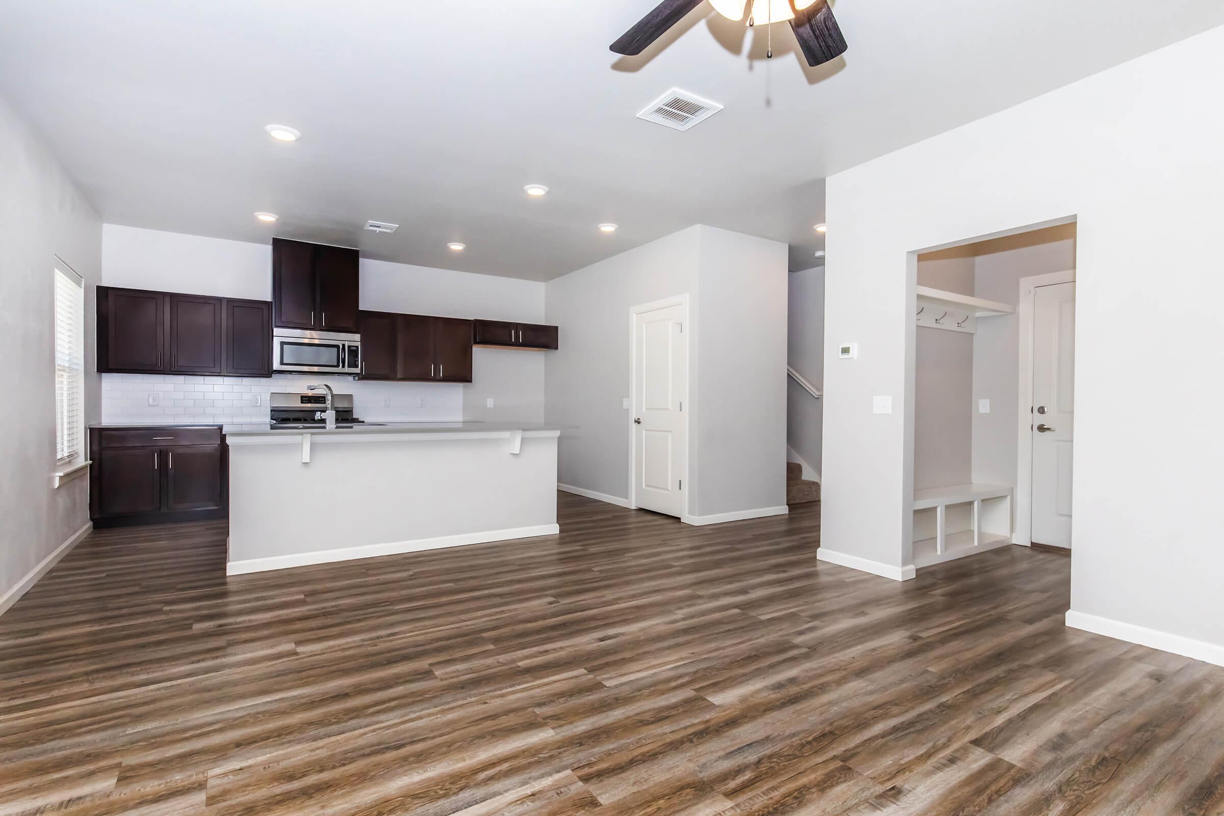 A modern, open-concept living area with hardwood-style flooring. The space features a kitchen with dark wood cabinets, stainless steel appliances, and a breakfast bar. There's a small entryway with a coat rack to the right, and white walls create a bright and spacious atmosphere. Ceiling fans add a contemporary touch.