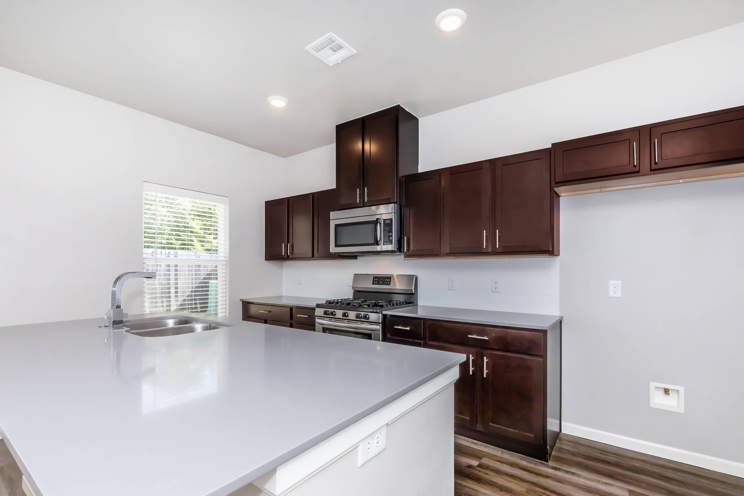 Modern kitchen featuring dark wooden cabinets, stainless steel appliances including a microwave and gas stove, a light-colored countertop, and a sink. Bright natural light comes in from a window, with a neutral color palette and wood-style flooring.