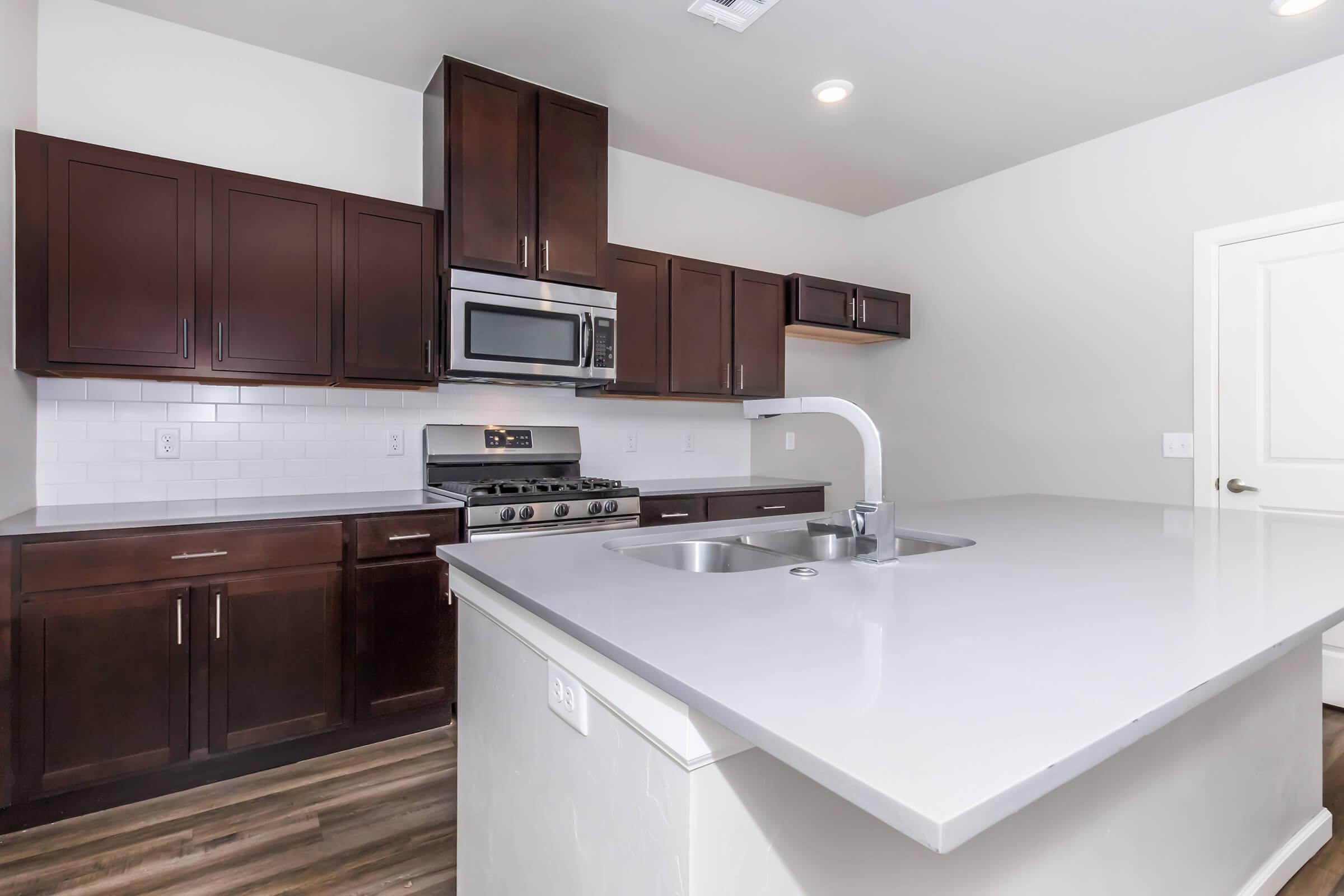 Modern kitchen featuring dark wood cabinets, stainless steel appliances including a microwave and gas stove, a double sink on a light countertop, and a neutral wall color, enhancing the bright and spacious atmosphere.