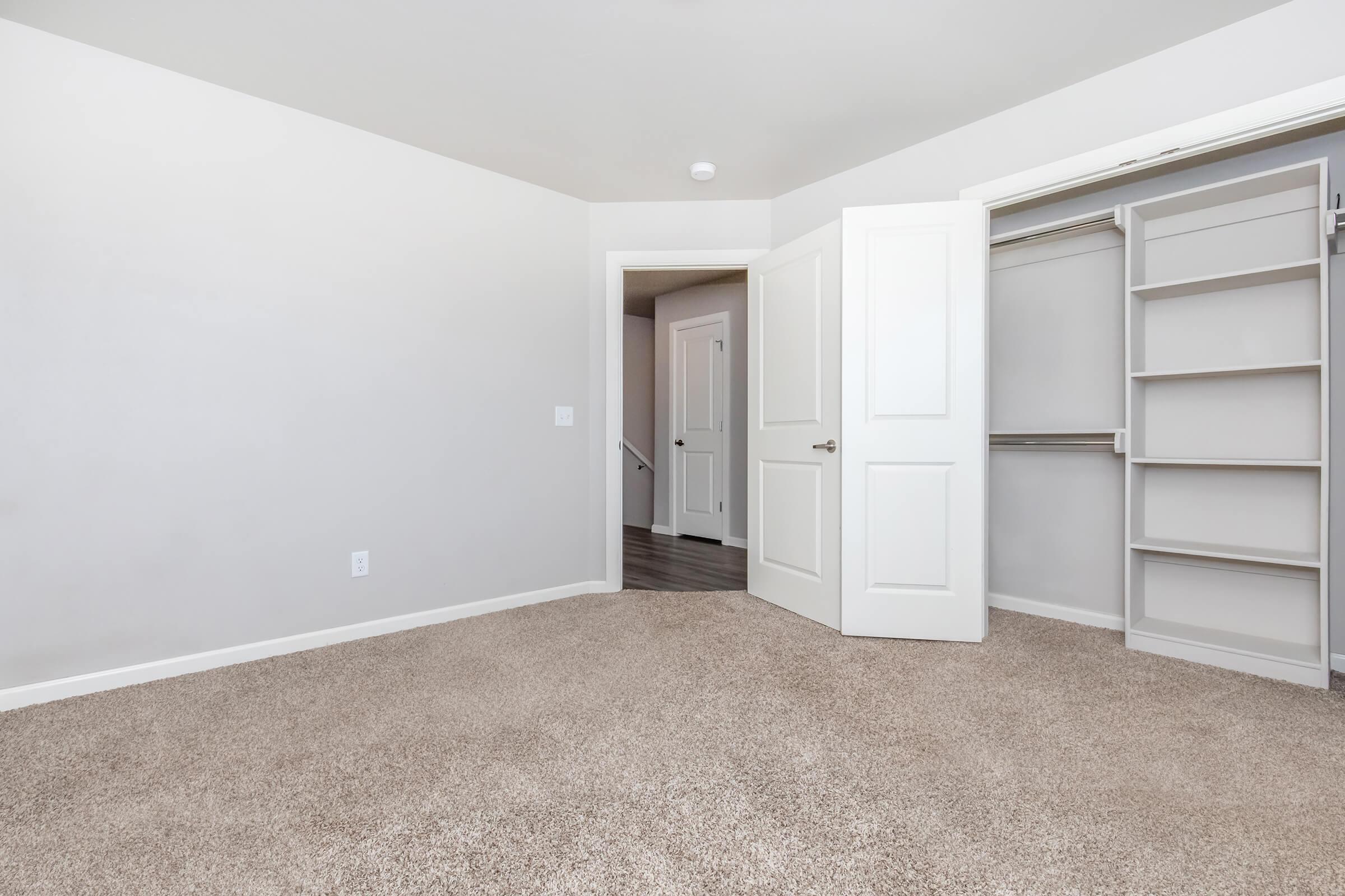 A spacious, empty bedroom featuring light-colored walls and plush carpet. An open closet with shelving is on one side, while a door leads to another room or hallway. The overall atmosphere is bright and uncluttered, ideal for personalization.