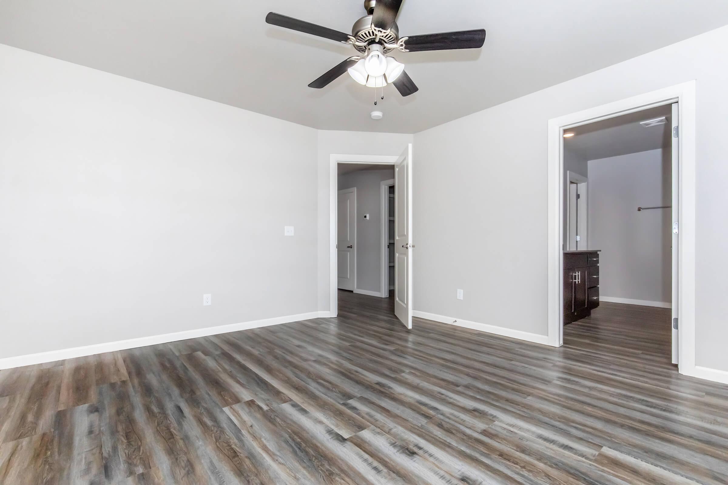 A spacious, empty room with light gray walls and a ceiling fan. The floor features gray-brown laminate planks. There are two doorways leading to other areas, one visible closet with a dark wooden cabinet. Natural light illuminates the room, creating a clean and modern appearance.