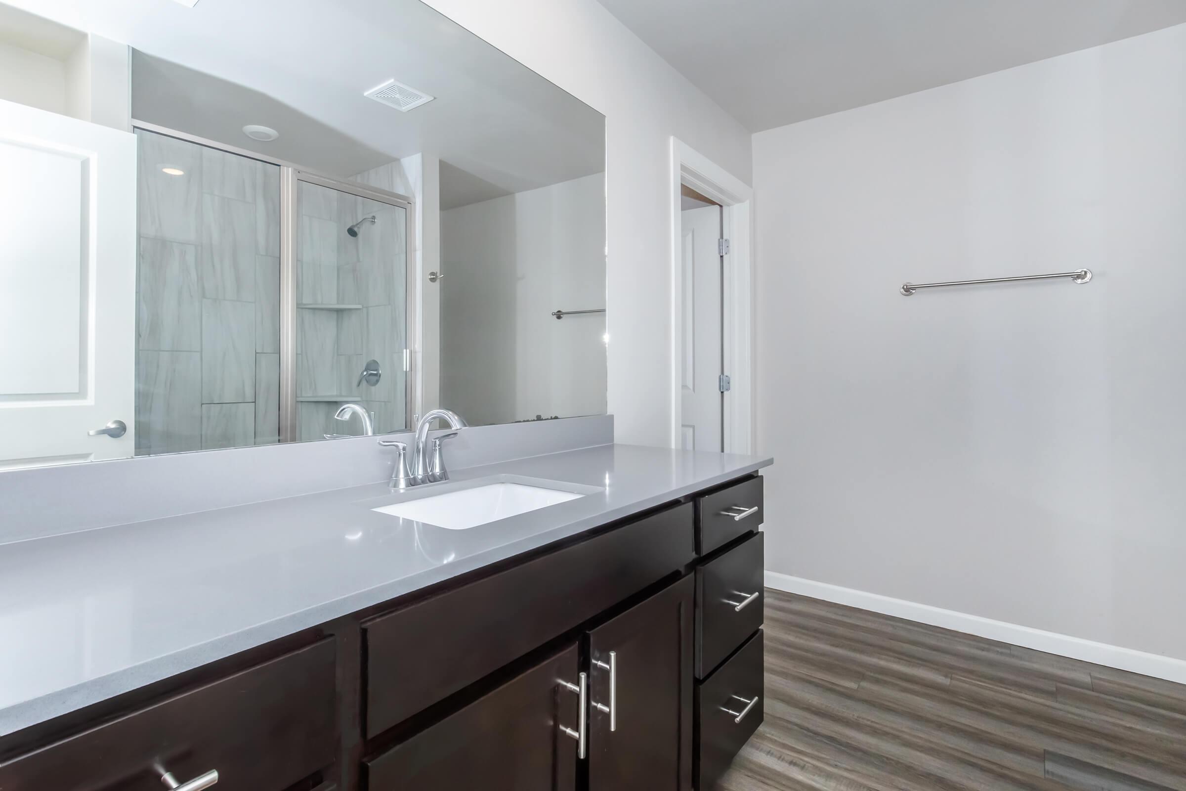 Modern bathroom featuring a double vanity with a sleek gray countertop and a single sink. The space has a large mirror above the vanity, white walls, and wood-like flooring. A glass shower is visible in the background, and there is a towel rack on the wall.