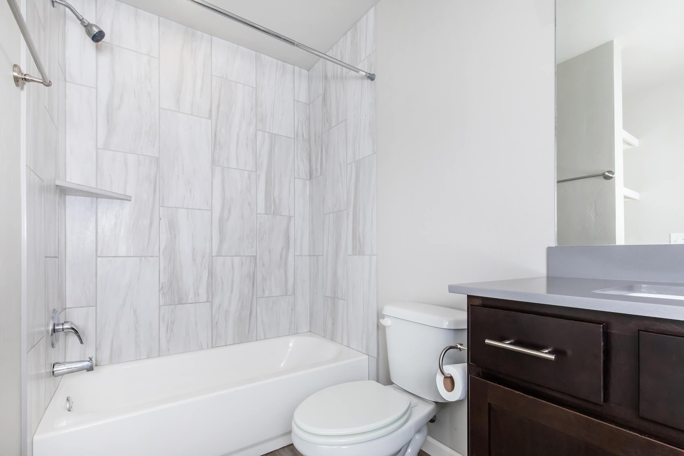 Modern bathroom featuring a clean design with a bathtub and shower combination, white marble-style tiles on the walls, a dark wood vanity with a gray countertop, and a toilet. Natural light brightens the space, creating a fresh and inviting atmosphere.