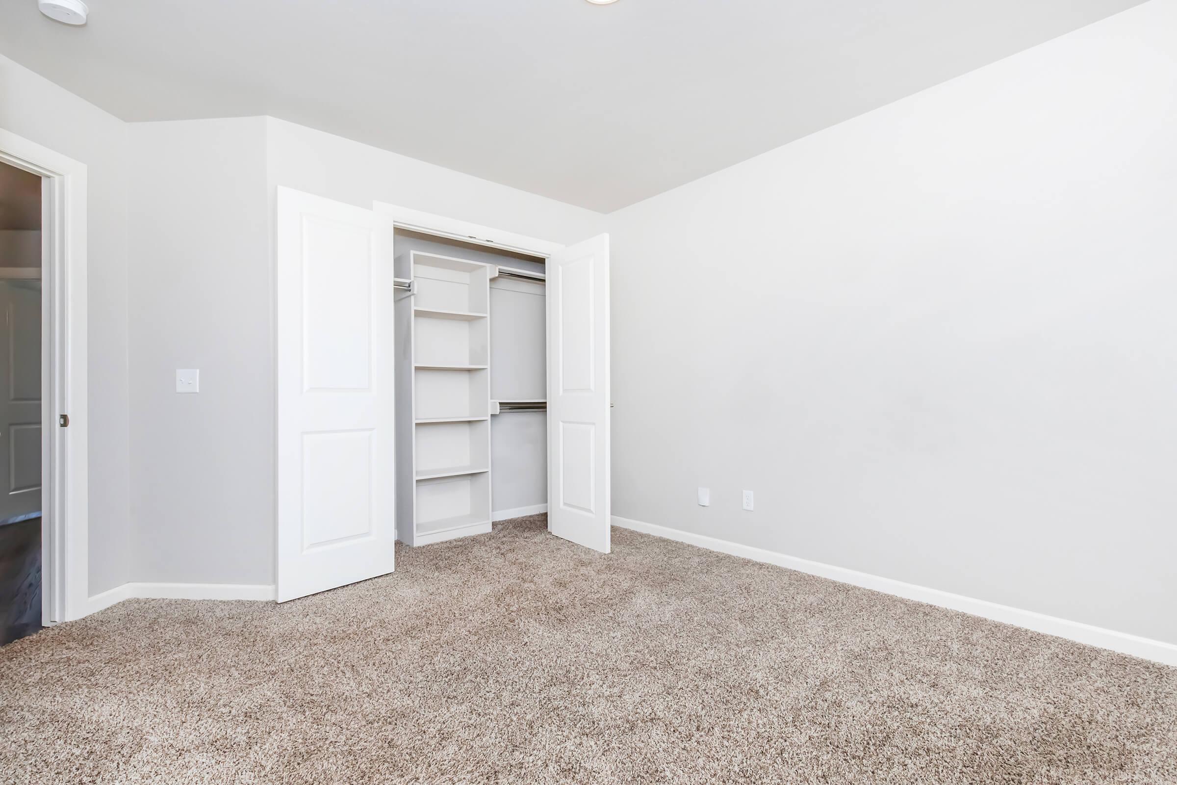 A clean, empty bedroom with light beige carpet. The walls are painted white, and there are two open closet doors revealing shelves inside. Natural light filters through, creating a bright atmosphere. The room has a minimalist feel, making it suitable for personalized decoration.