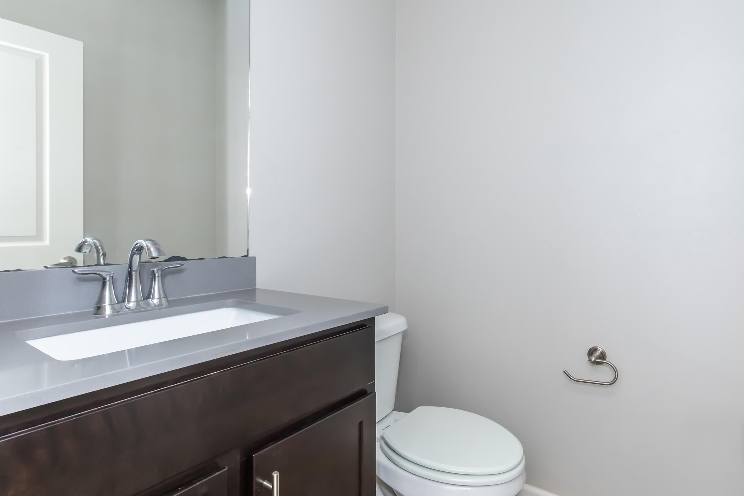 A bathroom featuring a modern sink with a sleek countertop, a mirror, and a white toilet. The walls are painted gray, and there is a towel bar on the wall beside the toilet.