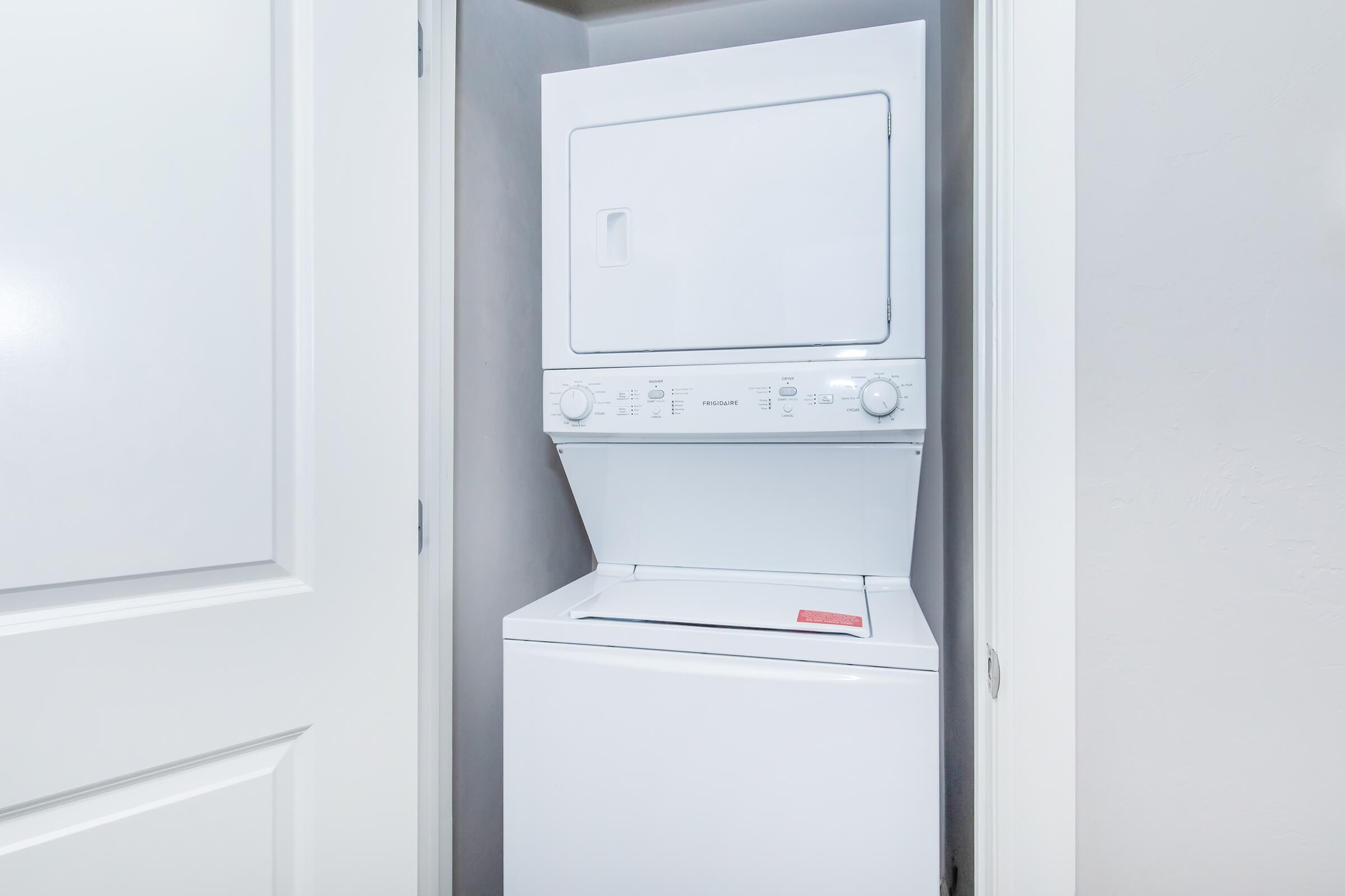 A compact white washer and dryer stacked in a small laundry closet, with a light-colored wall and door partially visible.