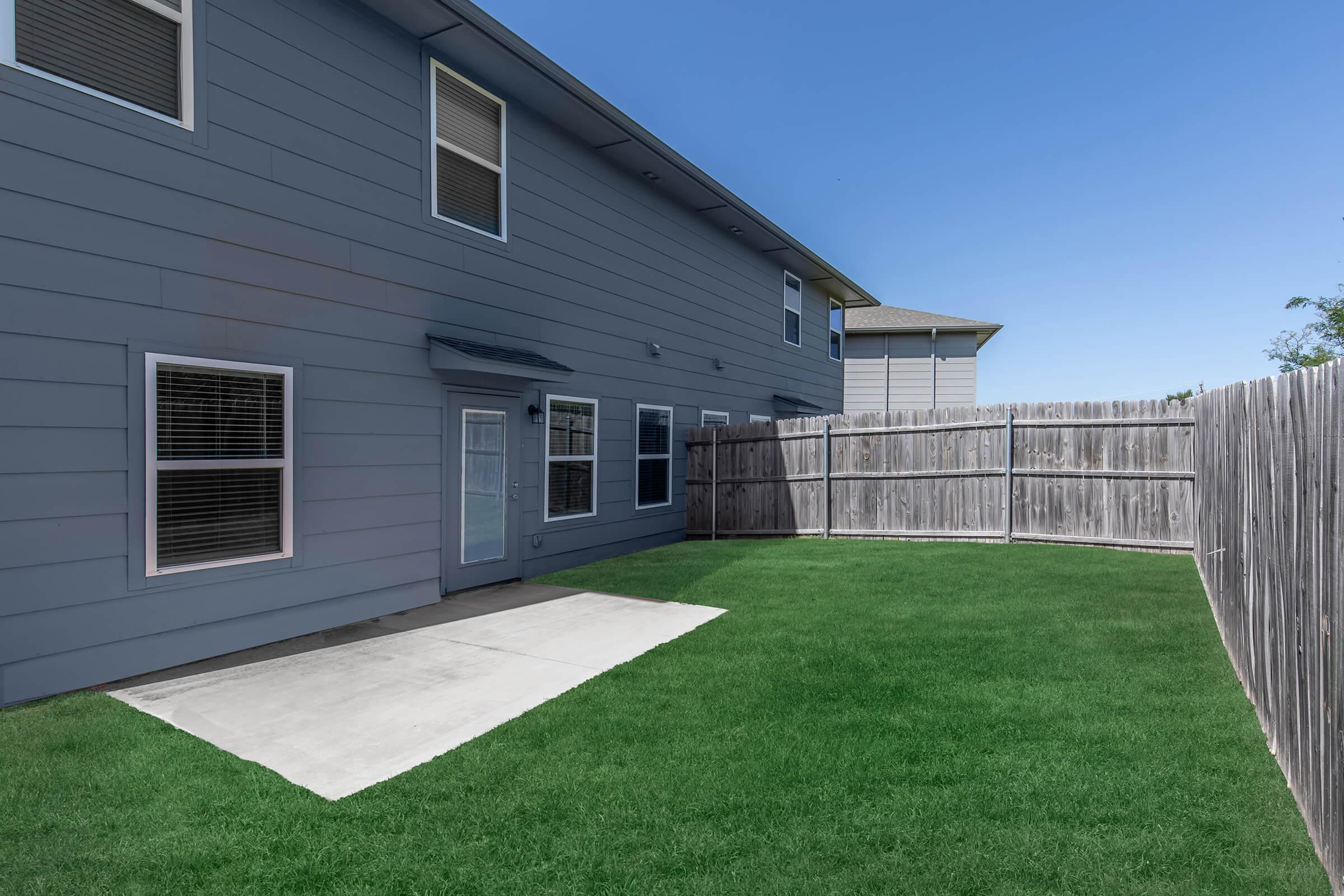 View of a backyard featuring a well-maintained green lawn, a concrete patio area, and a wooden fence. The side of a two-story gray house is visible, along with several windows that allow natural light into the home. The sky is clear and blue, indicating a sunny day.