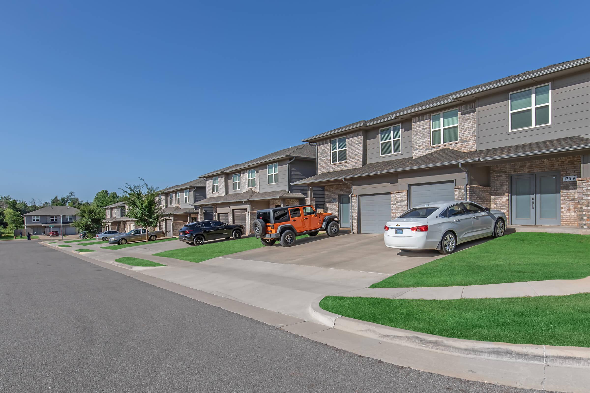 A quiet residential street featuring several houses with well-maintained landscaping. Vehicles are parked in driveways, including an orange Jeep and a white sedan. The sky is clear and blue, indicating a sunny day. The homes have a modern design with brick and siding exteriors.