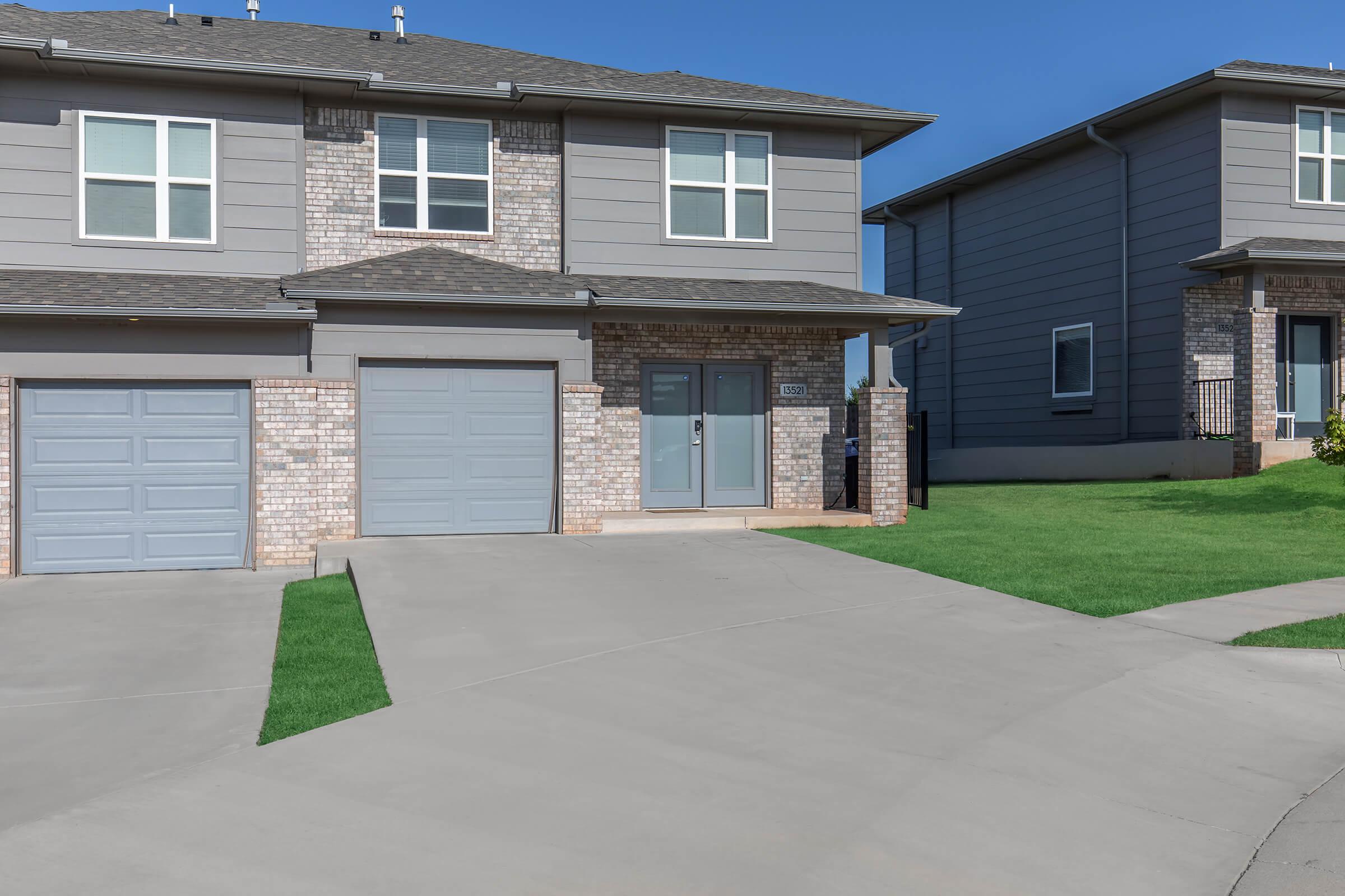 A modern two-story residential building with gray siding, featuring a brick exterior on the lower portion. The front yard is neatly landscaped with green grass, and there are two garages with light gray doors. The sky is clear and blue, enhancing the overall aesthetic of the property.