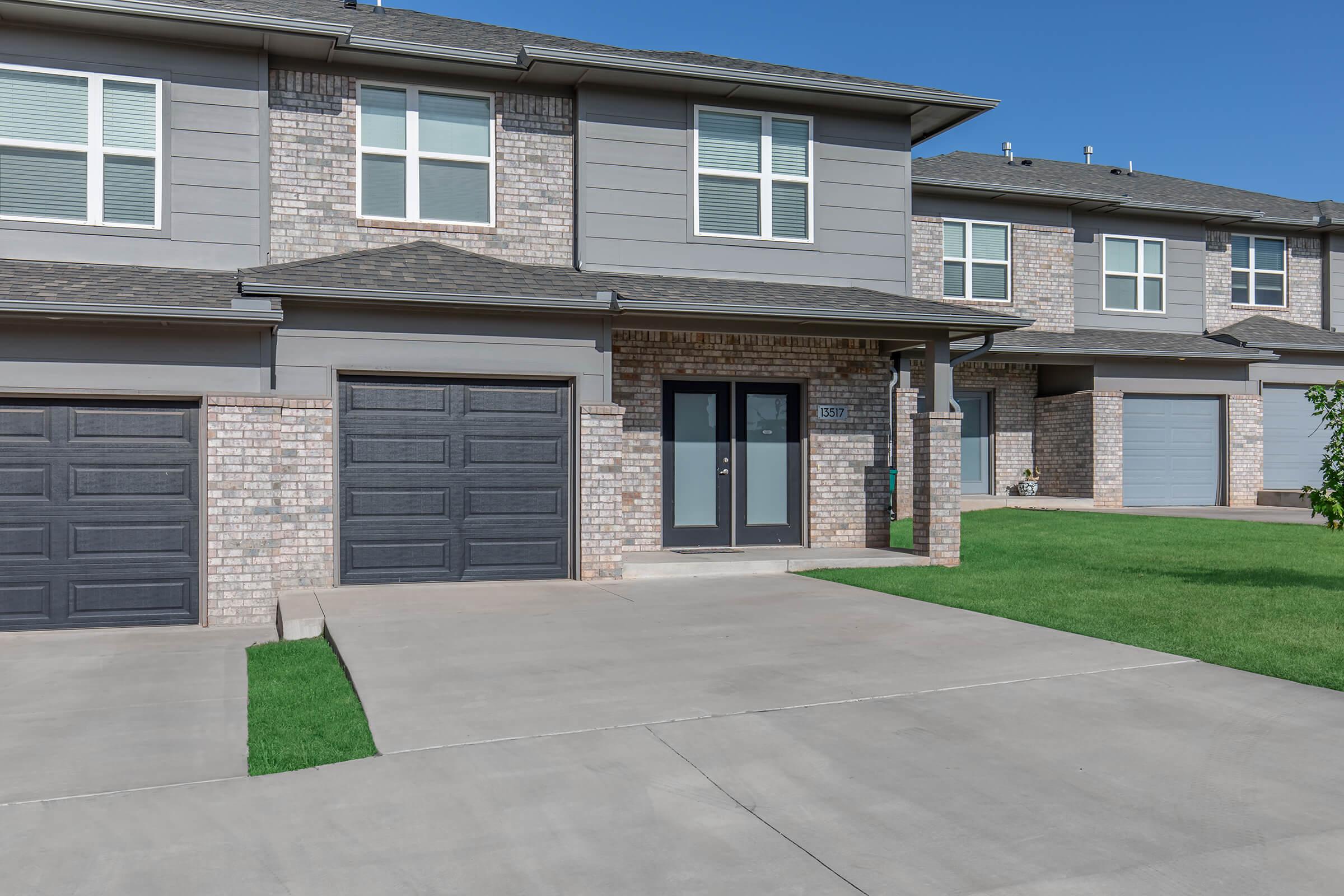 A modern two-story residential building featuring brick and gray siding, with a driveway and garage doors. The front has a small porch area and well-manicured green lawns on either side, under a clear blue sky.