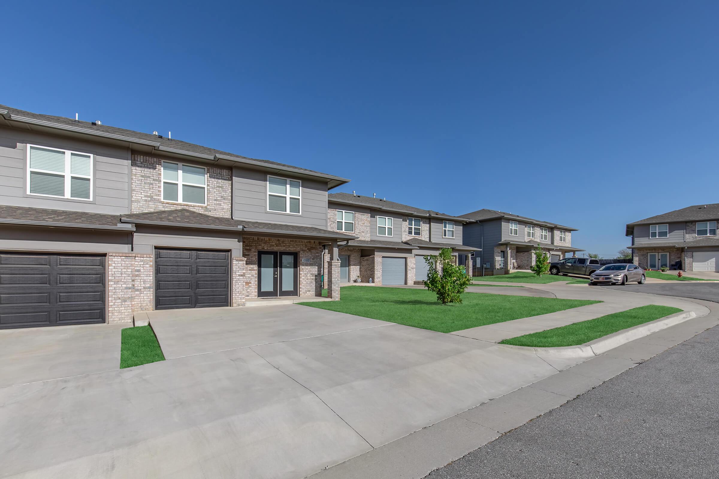 Row of modern townhouses with gray and brick exteriors, featuring one-car garages. The driveway is paved, and well-maintained green lawns are visible in front of the homes. Clear blue sky overhead, indicating a sunny day. The scene conveys a suburban residential area.