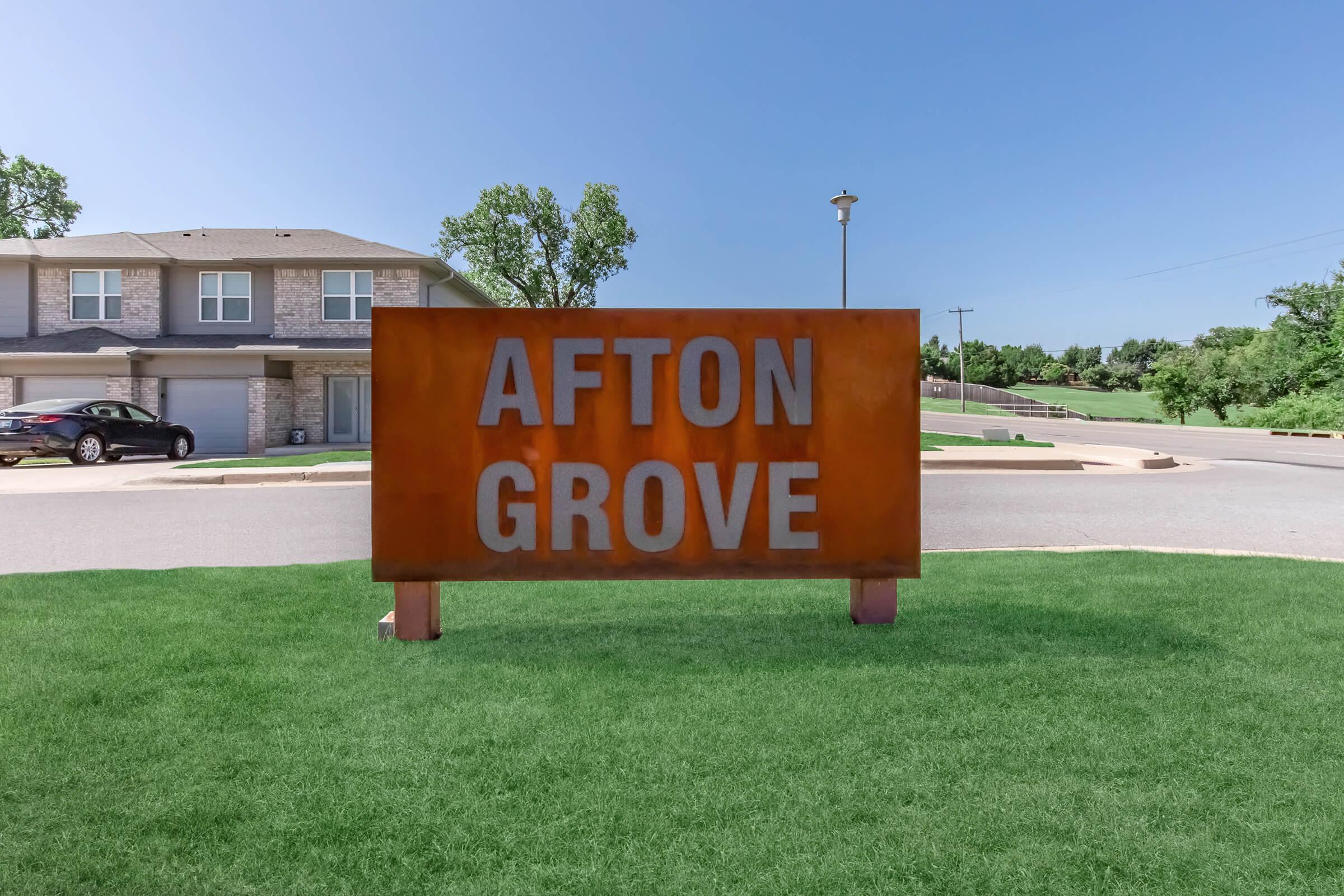 Afton Grove sign displayed prominently on a grassy area, featuring large, bold letters against a rust-colored background. The surrounding landscape includes a clear blue sky and a few trees, with a road and residential buildings visible in the background.