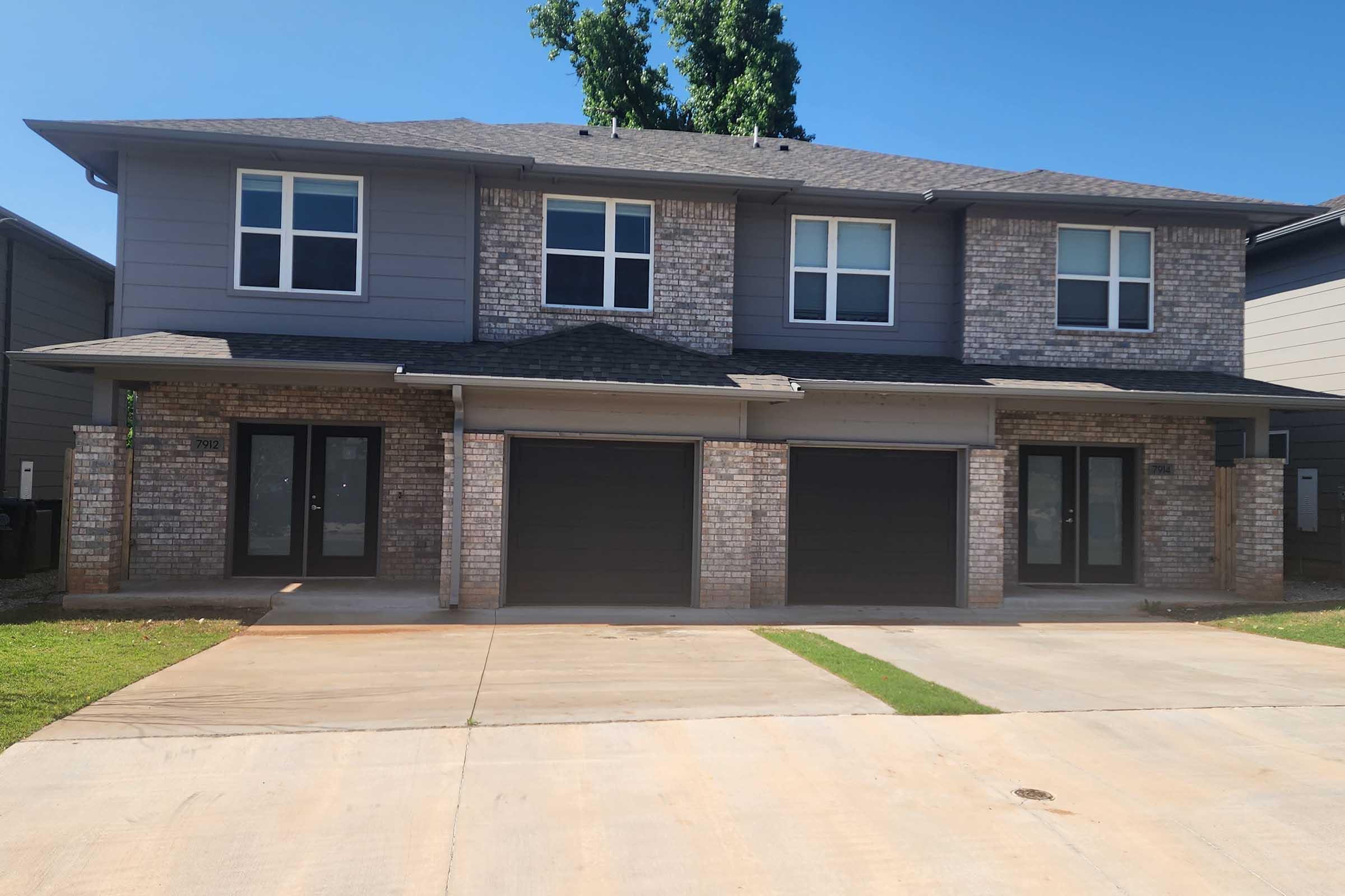 A modern two-story duplex featuring a brick and siding exterior, with three large windows on the upper floor. The lower floor has two garage doors and a concrete driveway. The property is surrounded by a well-maintained lawn and trees. The sky is clear and blue.
