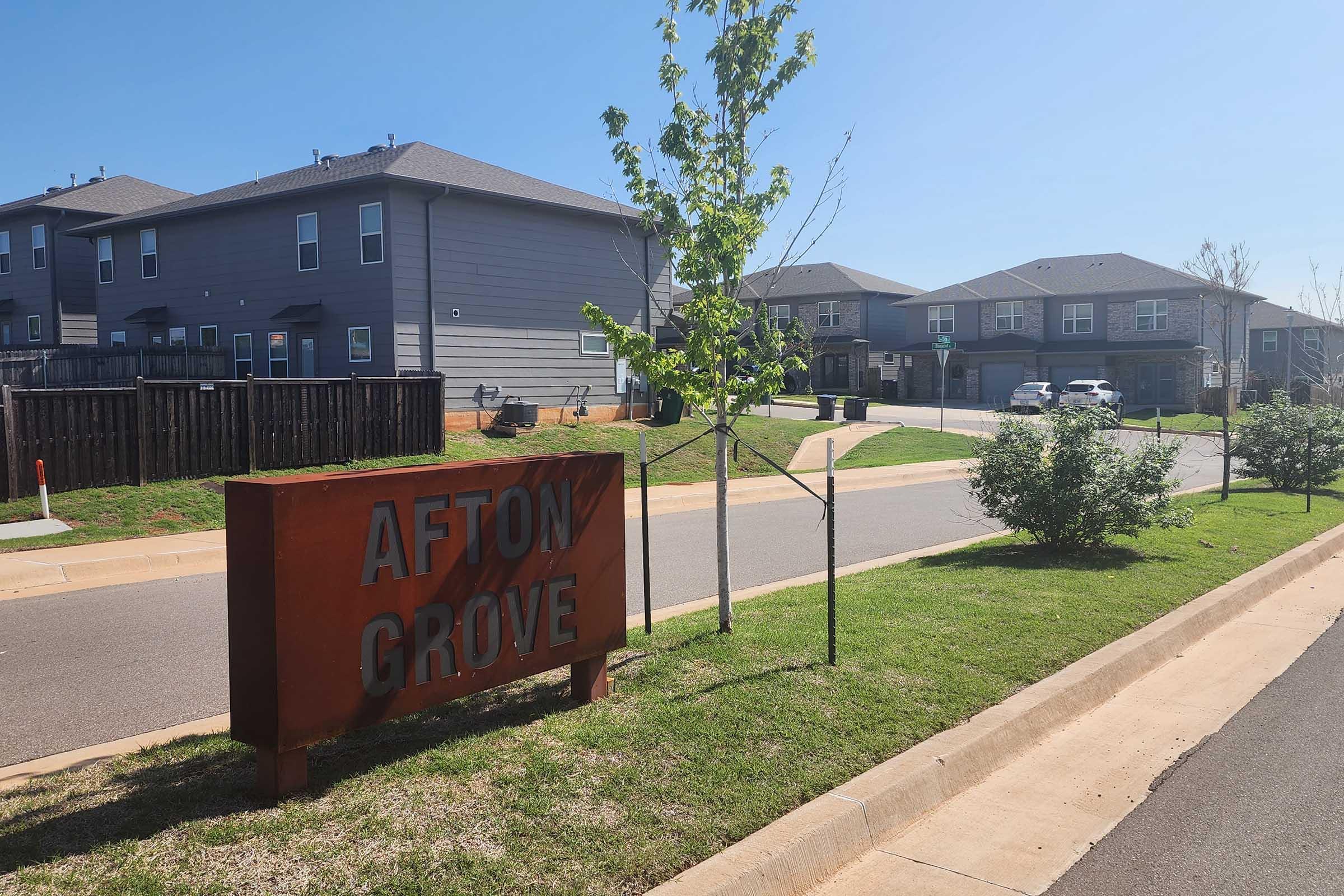 Afton Grove neighborhood sign displayed prominently alongside a neat, paved road. Residential buildings are visible in the background, with clear blue sky overhead and green landscaping including small trees and shrubs on either side of the street.
