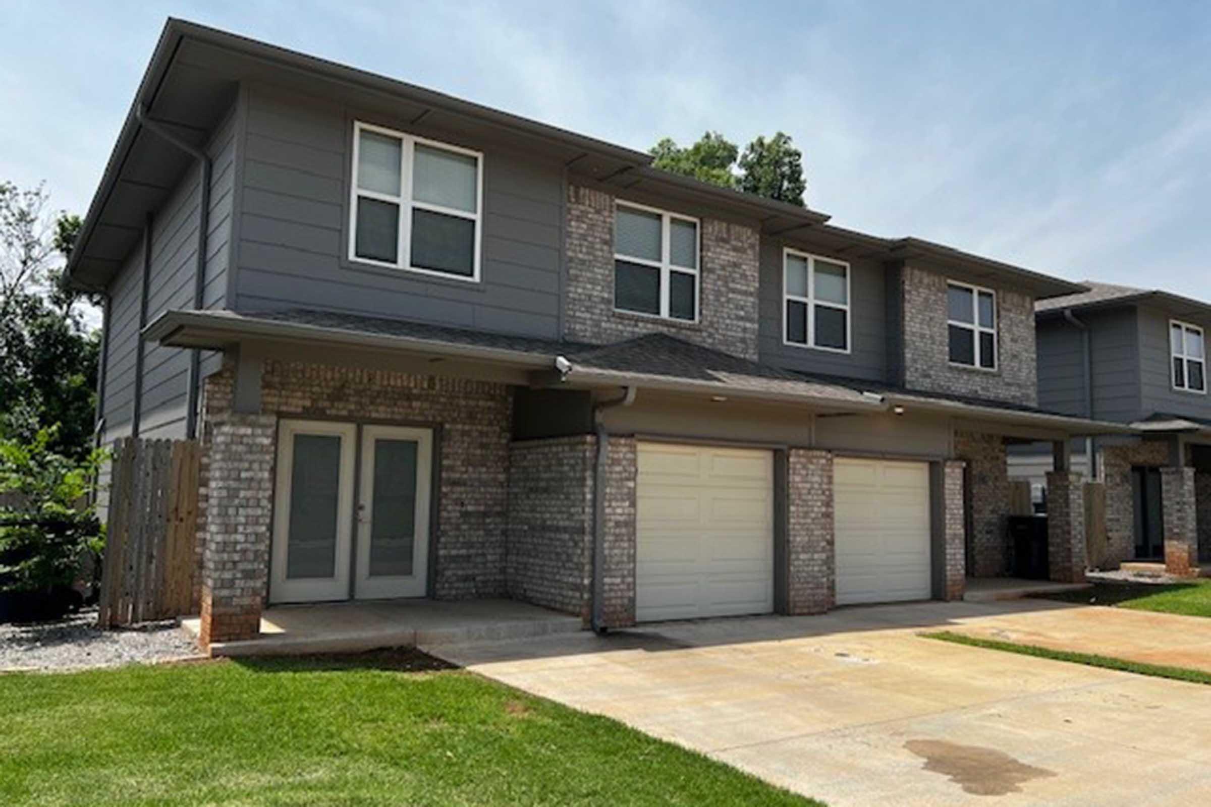 A modern two-story duplex with a brick and siding exterior. The front features two garage doors, a small porch area, and a driveway. Green grass and a tree are visible in the surroundings, with a clear blue sky in the background.