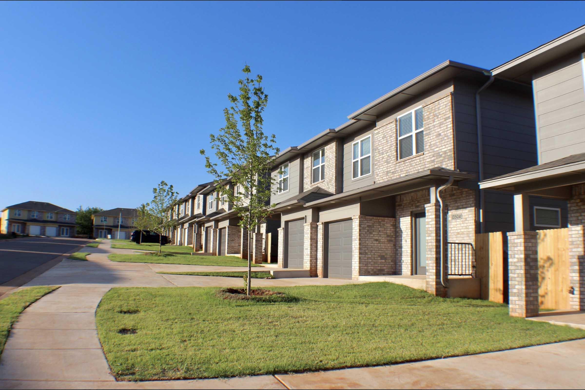 Row of modern townhouses with two stories, featuring a mix of brick and siding exteriors. Neatly trimmed lawns and small trees line the pathway. Clear blue sky in the background, creating a bright and welcoming atmosphere.