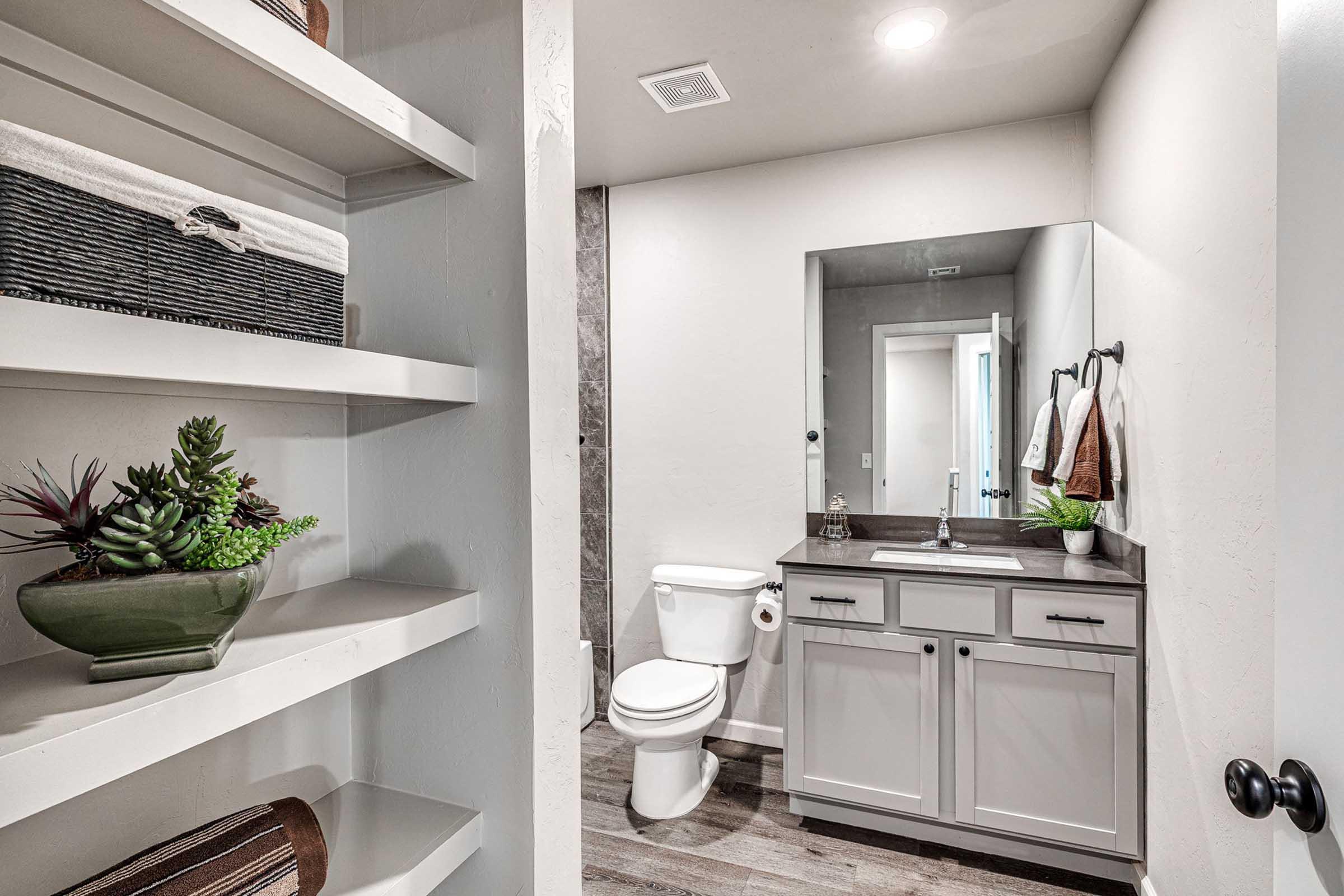 A modern bathroom featuring a toilet, a sink with a dark countertop, and a large mirror. White shelves hold decorative items and plants. The walls are light-colored, and there is a towel rack with two towels. The flooring has a wood-like appearance, enhancing the contemporary design.