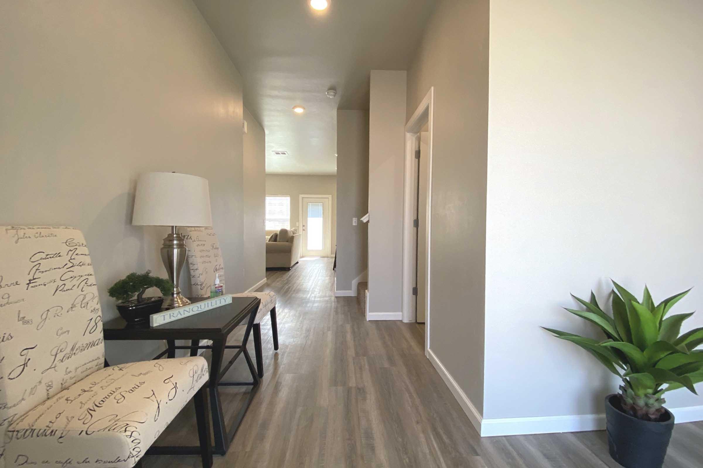 A well-lit hallway with light-colored walls and wooden flooring. Two stylish chairs with patterned upholstery are positioned beside a lamp on a small table. A potted plant adds a touch of greenery at the end of the hall, leading towards a sunlit room visible in the distance.