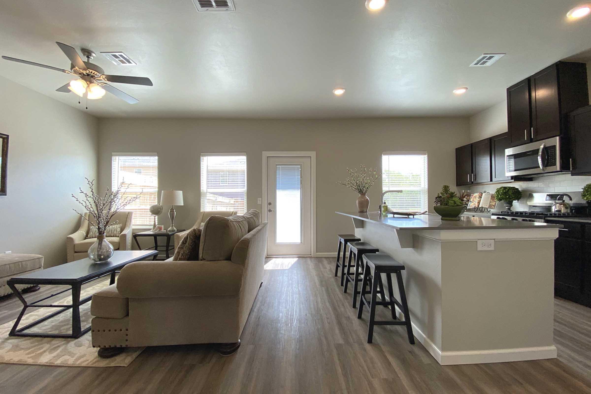 Interior view of a modern living room and kitchen area. The room features a comfortable sofa, a coffee table, and a dining area with bar stools. Natural light streams in through large windows, and a door leads outside. The kitchen has dark cabinetry and stainless steel appliances, complemented by decorative plants.