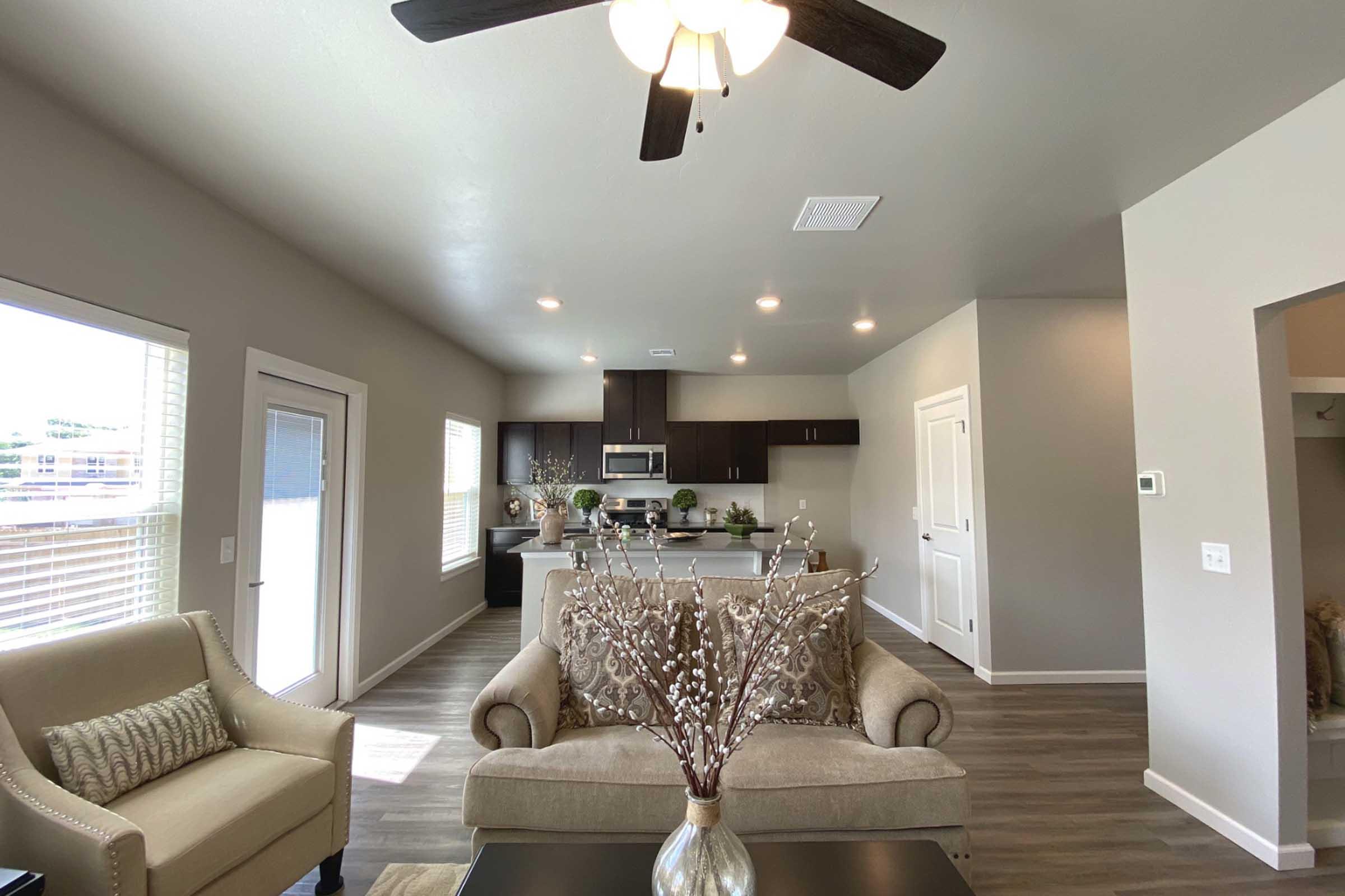 A cozy living room with a beige sofa and decorative branches in a vase. Natural light streams through the windows, illuminating a modern kitchen with dark cabinetry in the background. A ceiling fan hangs overhead, and the space features light wood flooring and neutral-colored walls, creating an inviting atmosphere.